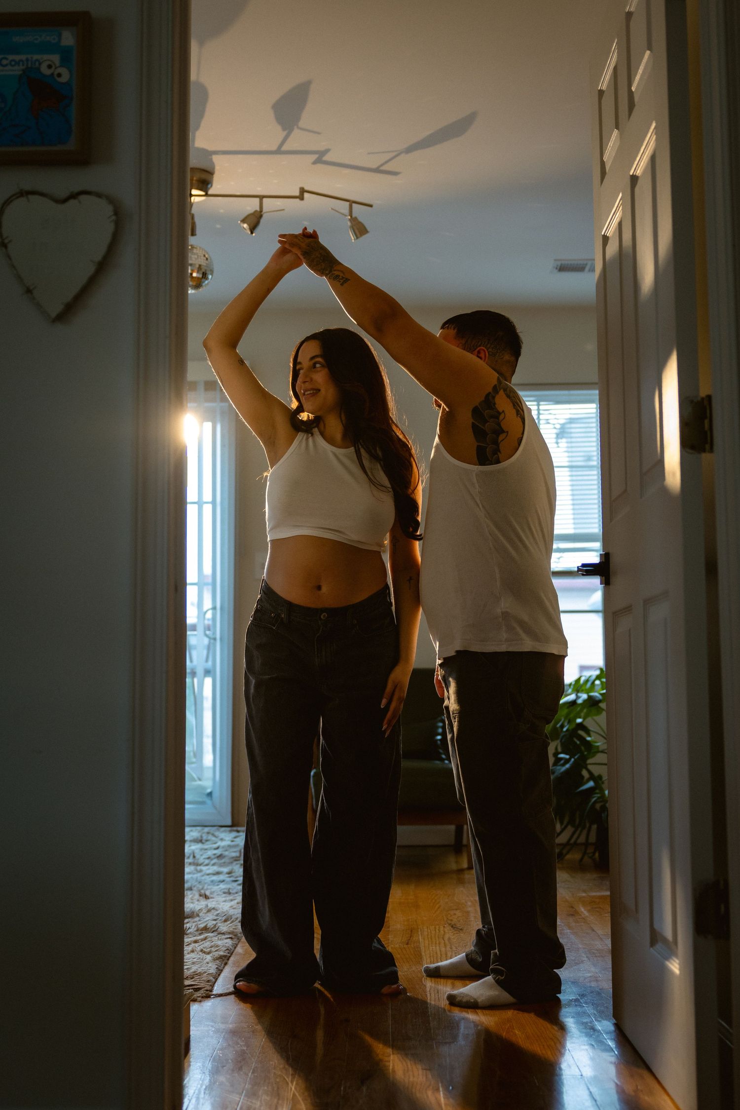 A couple dances together in a warmly lit home hallway, the man spinning the woman who smiles in a white crop top.