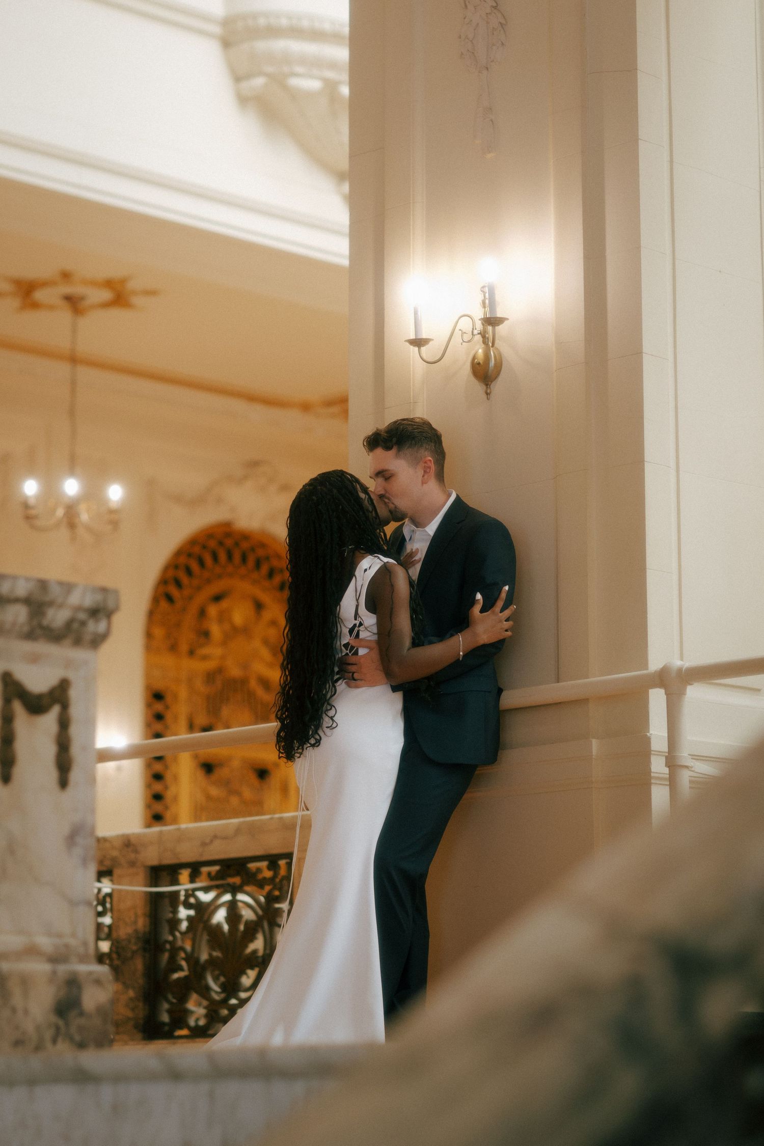 Interracial couple sharing a romantic moment in an elegant wedding venue with ornate decor and warm lighting.