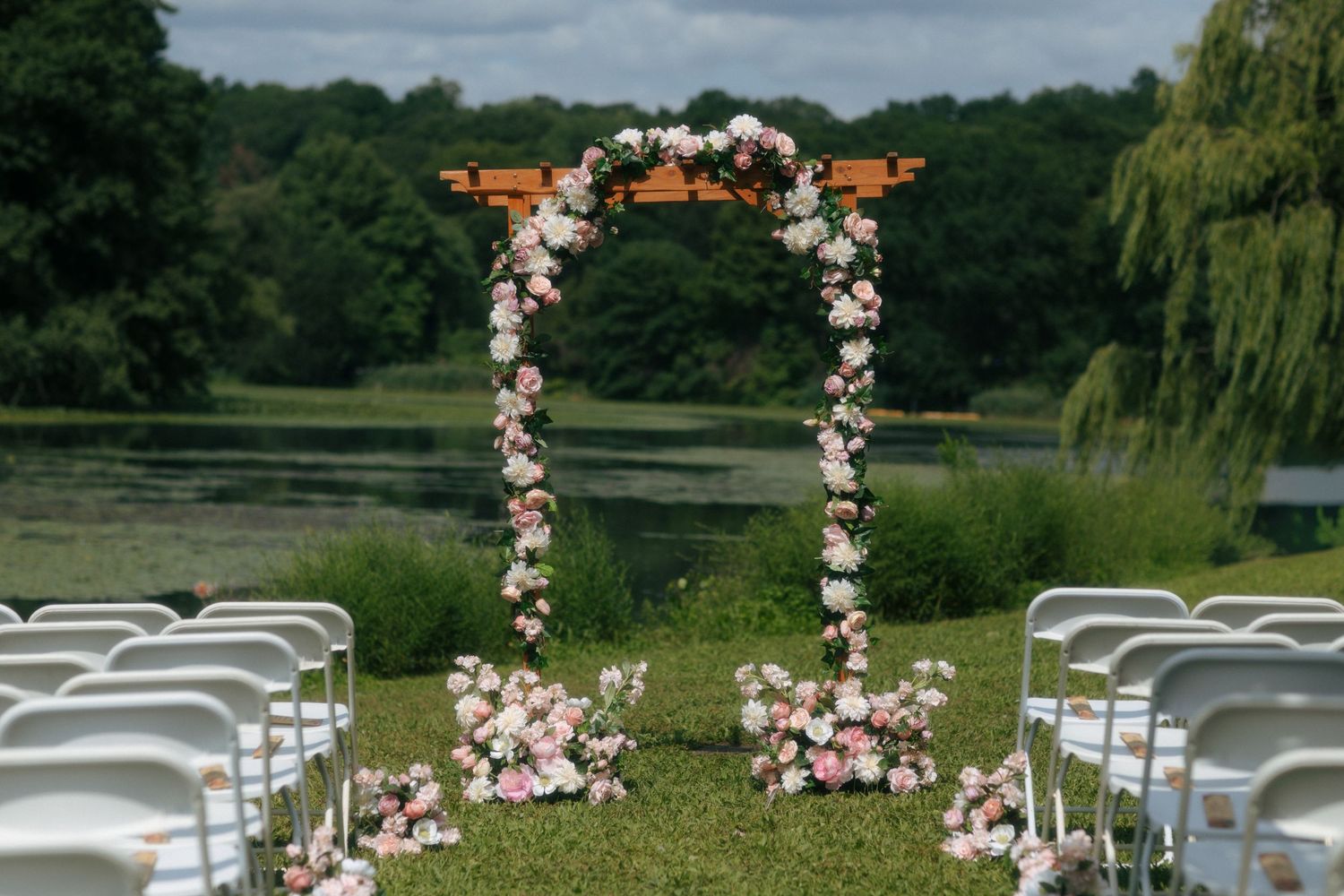 Outdoor wedding ceremony setup with floral arch of pink roses beside a serene lake.