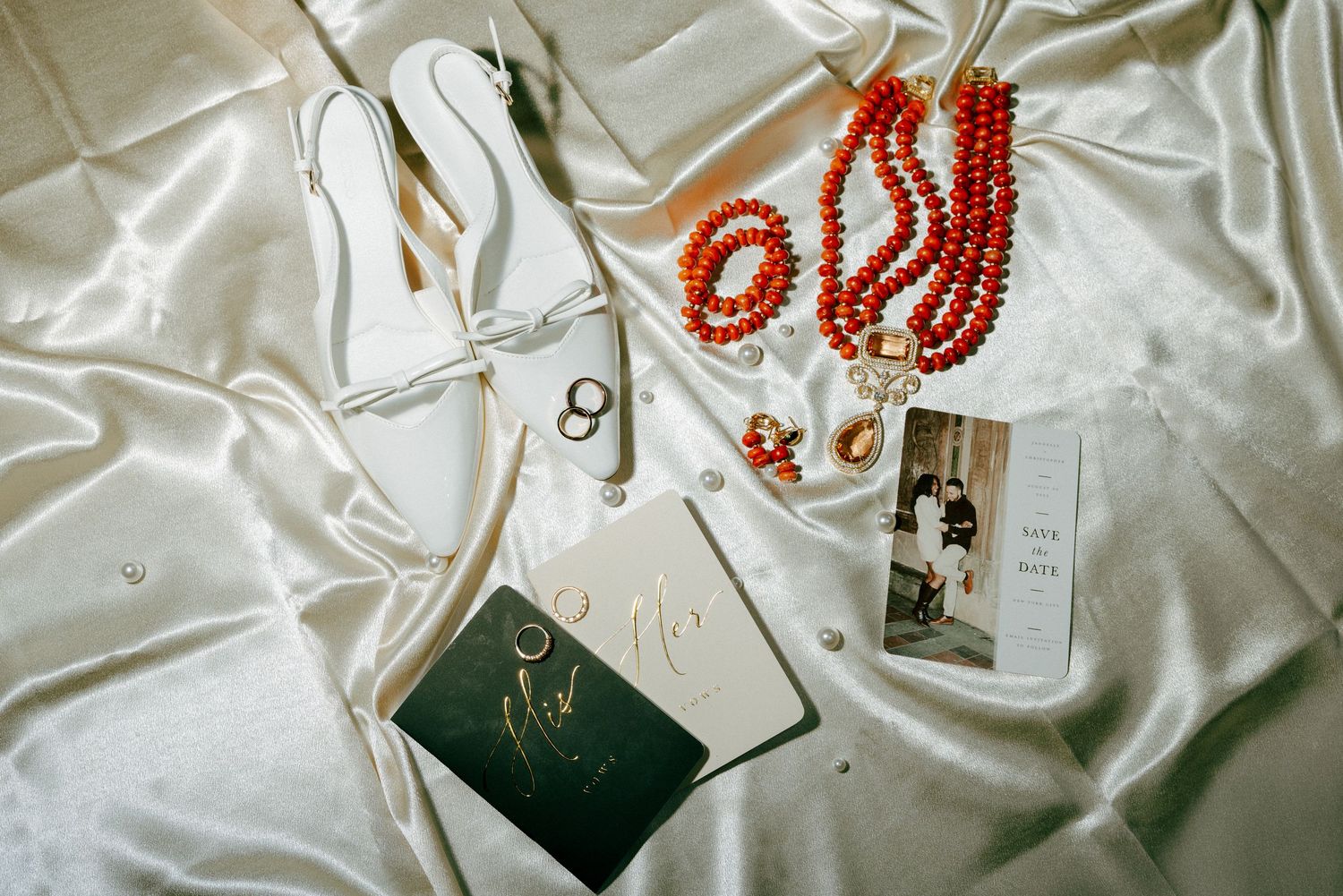 Bridal flat shoes, red beaded jewelry, vow books, and save the date on white satin.