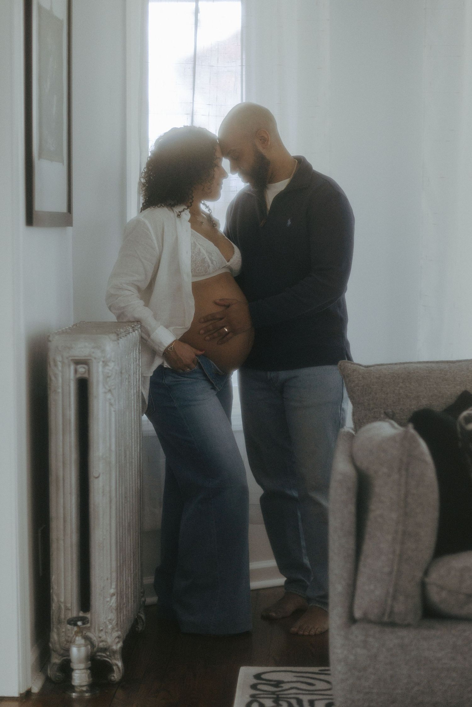 Pregnant couple sharing a tender moment by a window, woman showing baby bump in white top and jeans.