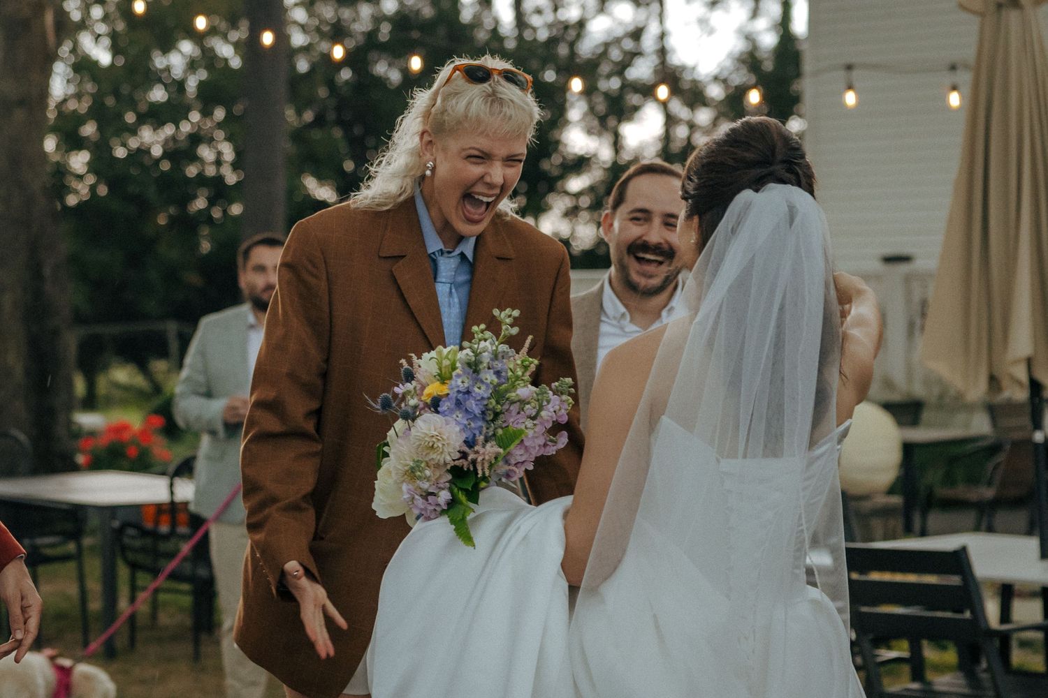 Joyful wedding moment with bride in white gown and veil, guest in brown coat holding colorful bouquet, laughing outdoors.