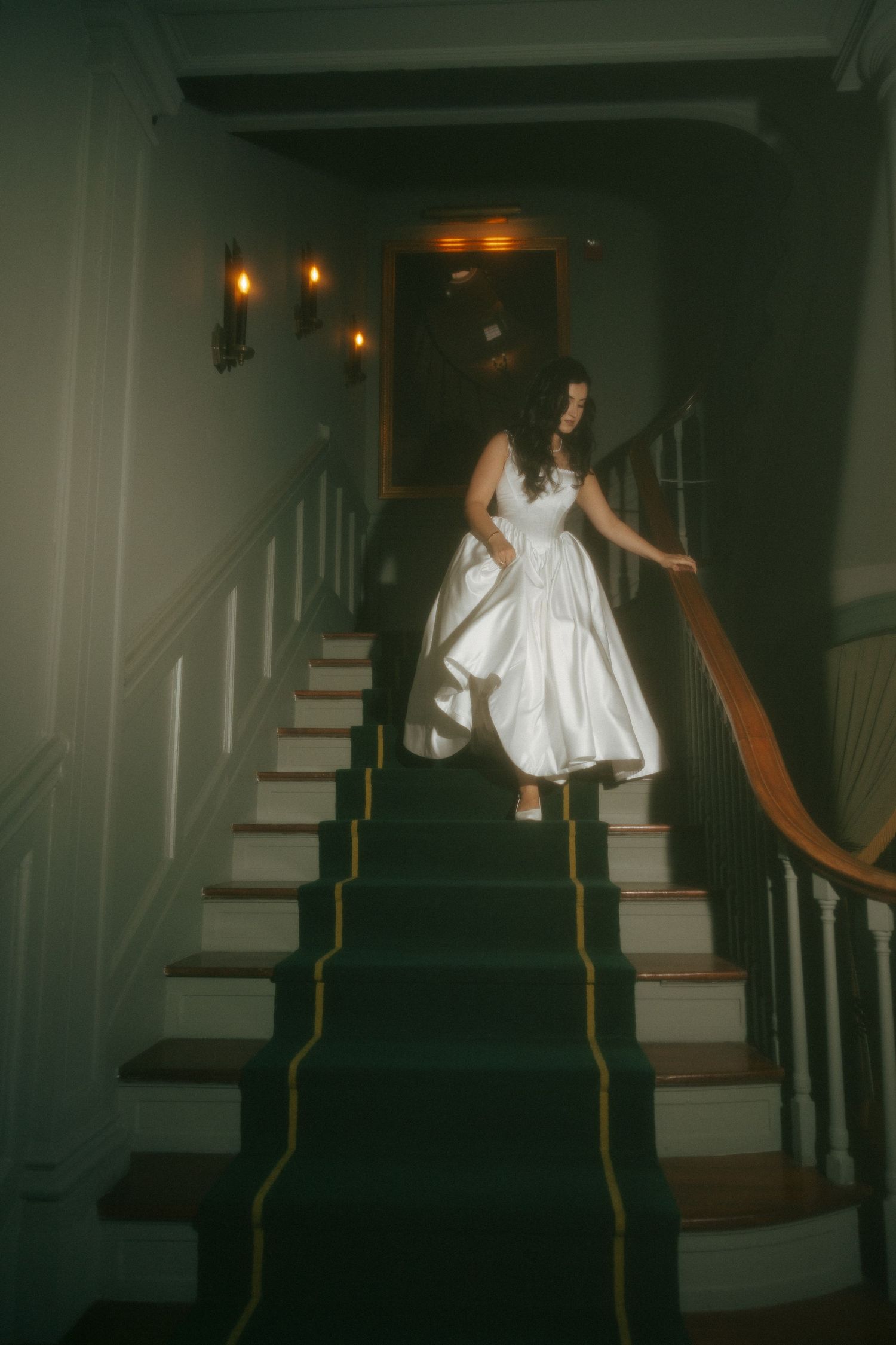 Bride in white satin wedding dress descending dark staircase with green carpet and wooden banister.