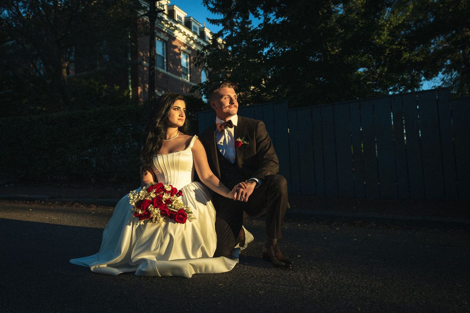 Bride and groom pose together outdoors at golden hour, she holds red bouquet in white gown, he wears a gray suit.