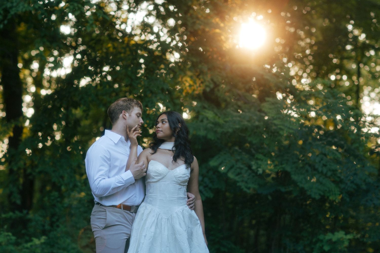 Bride and groom share a romantic moment in a sunlit forest, golden light streaming through lush green trees.