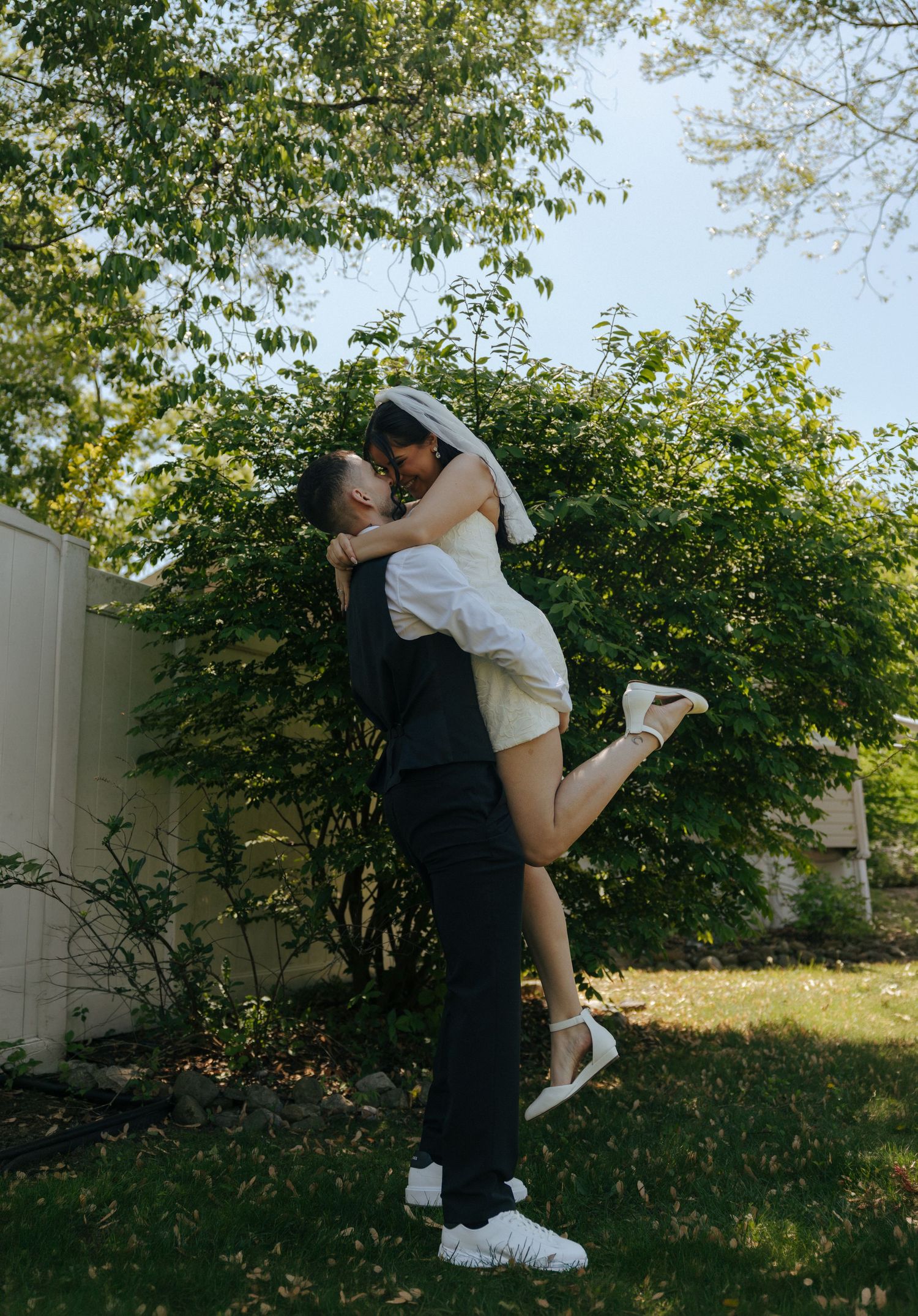 A groom lifts his bride in a white dress and veil outdoors, sharing a romantic kiss surrounded by lush green trees.