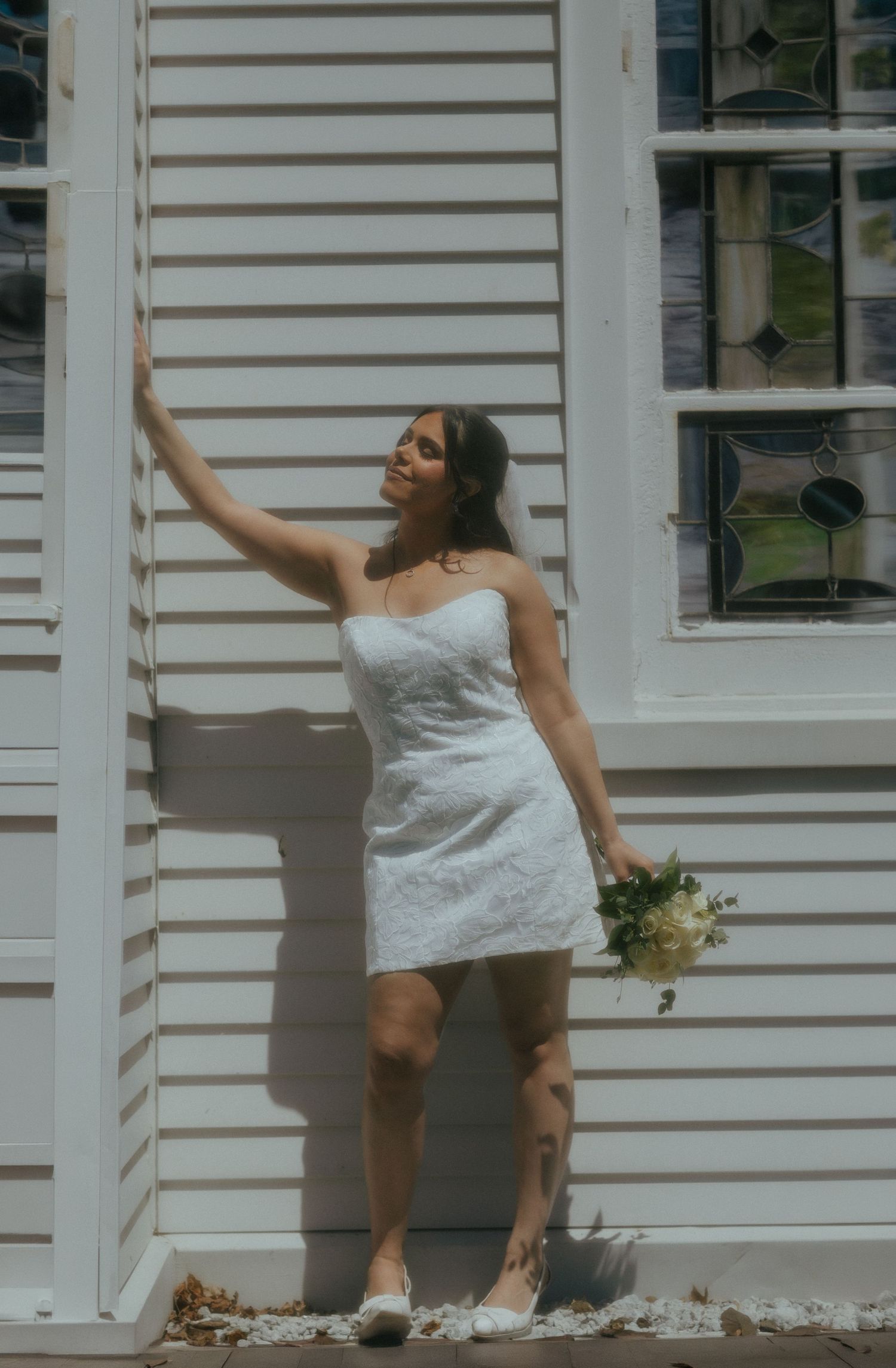 Bride in short white lace strapless dress holding bouquet, posing against white clapboard church wall in sunlight.