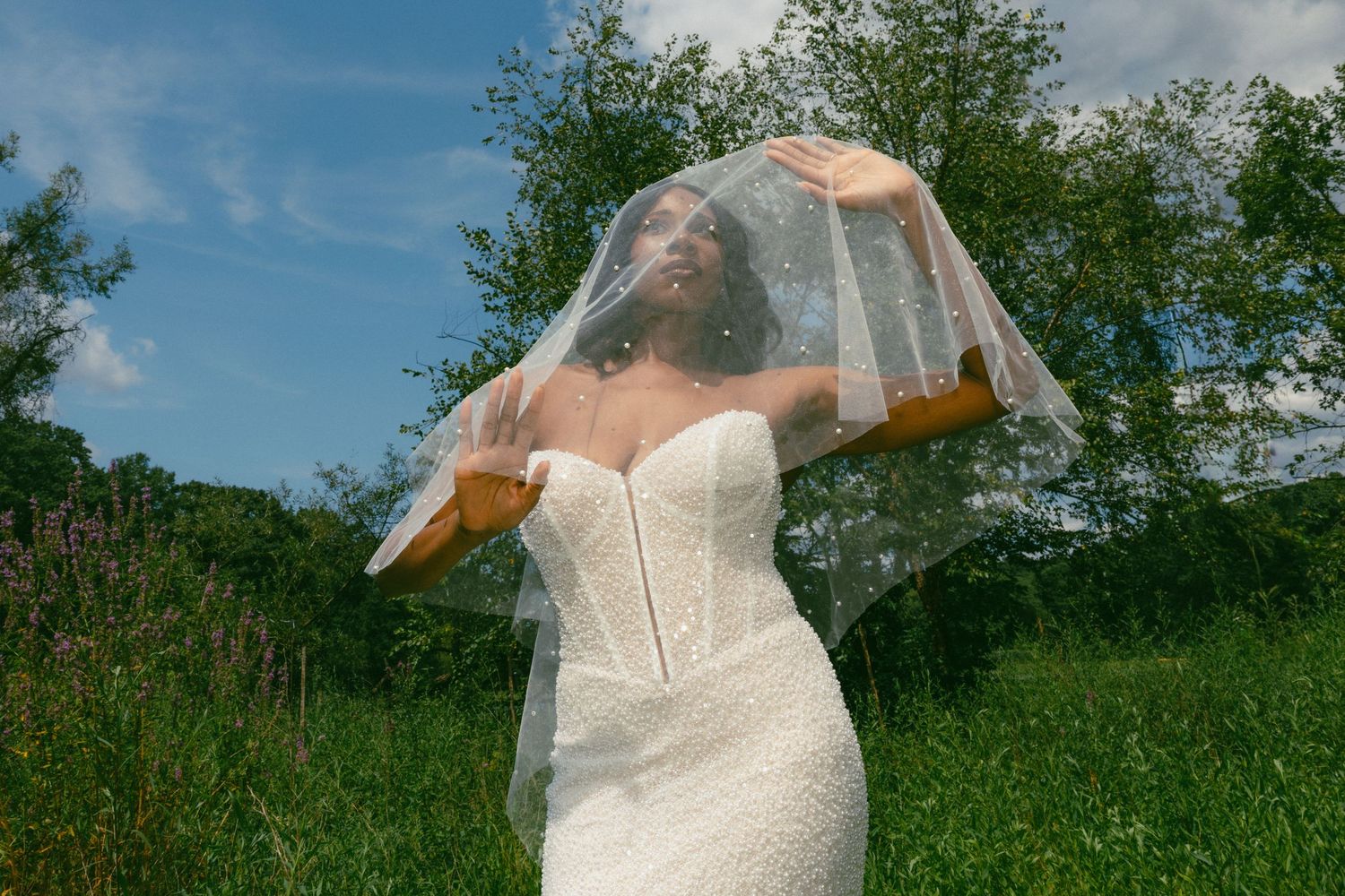 Bride in strapless white gown lifting her sparkling veil against a blue sky.
