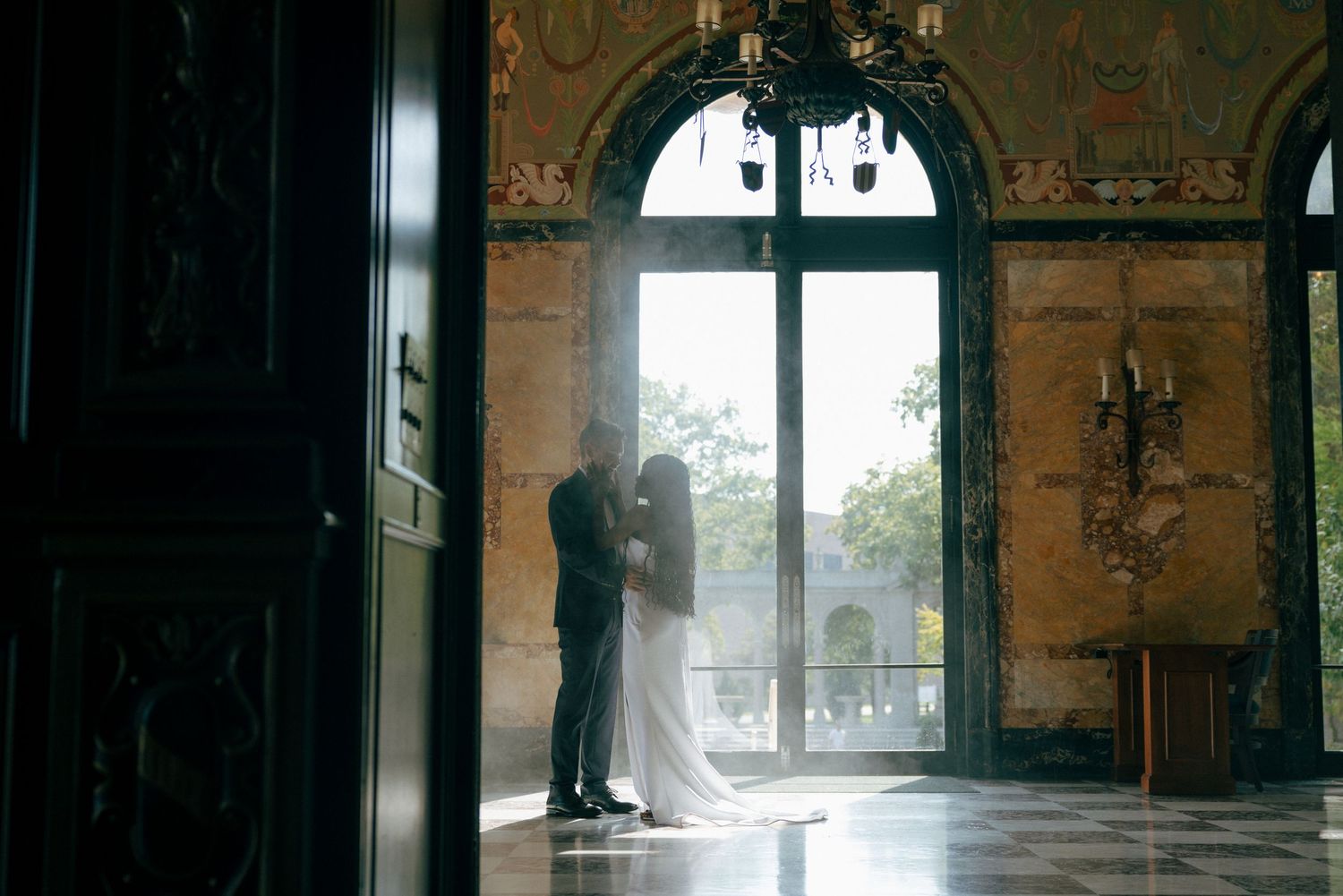 Bride and groom silhouetted in romantic embrace before grand arched window in ornate wedding venue.