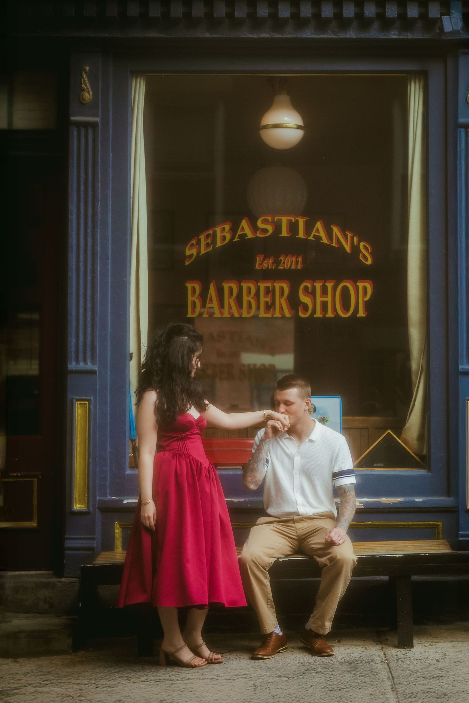 Couple posing outside Sebastian's Barber Shop, woman in red dress, man kissing her hand on a bench.