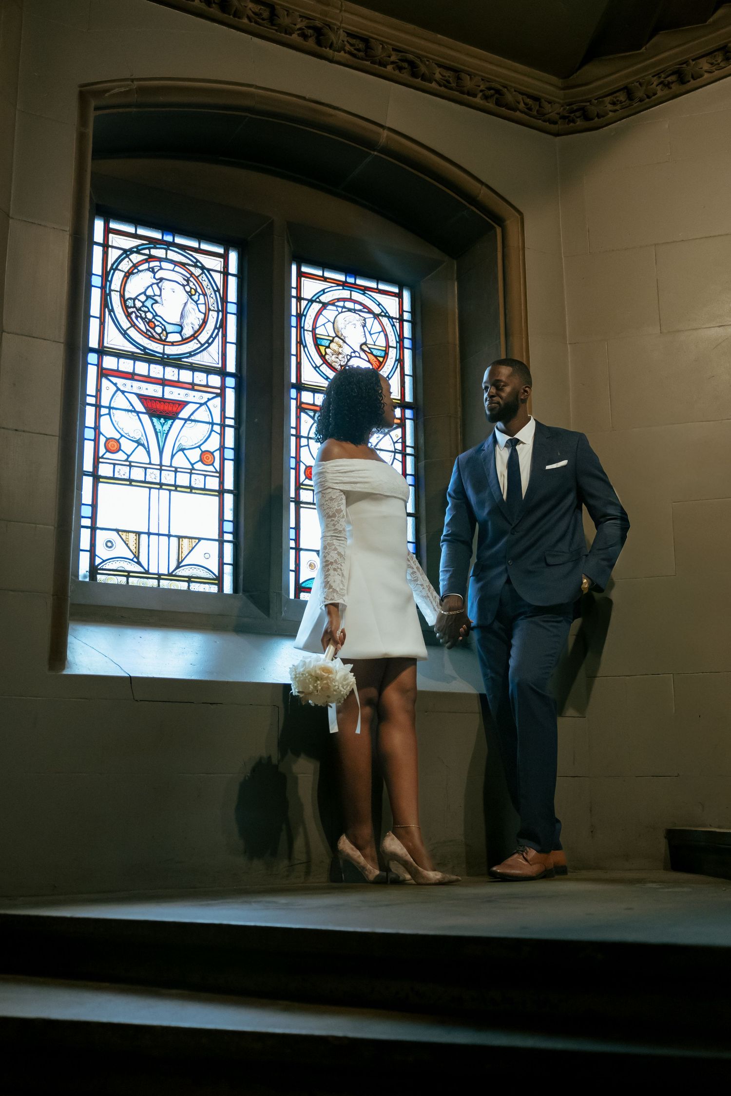 Elegant Black couple in wedding attire standing by stained glass church window, bride holding bouquet.
