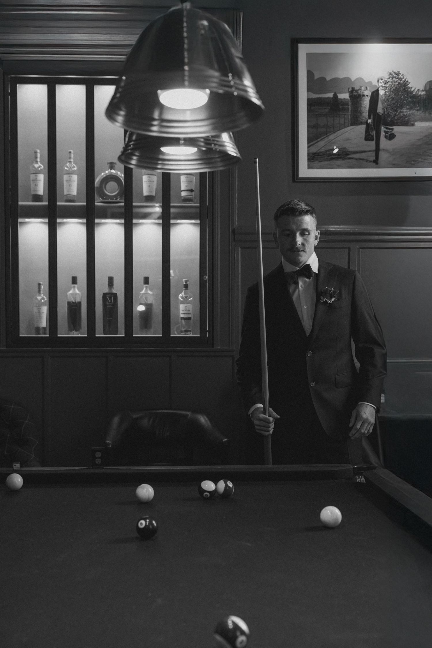 Groom in tuxedo holding pool cue beside billiard table in elegant game room with bar cabinet.
