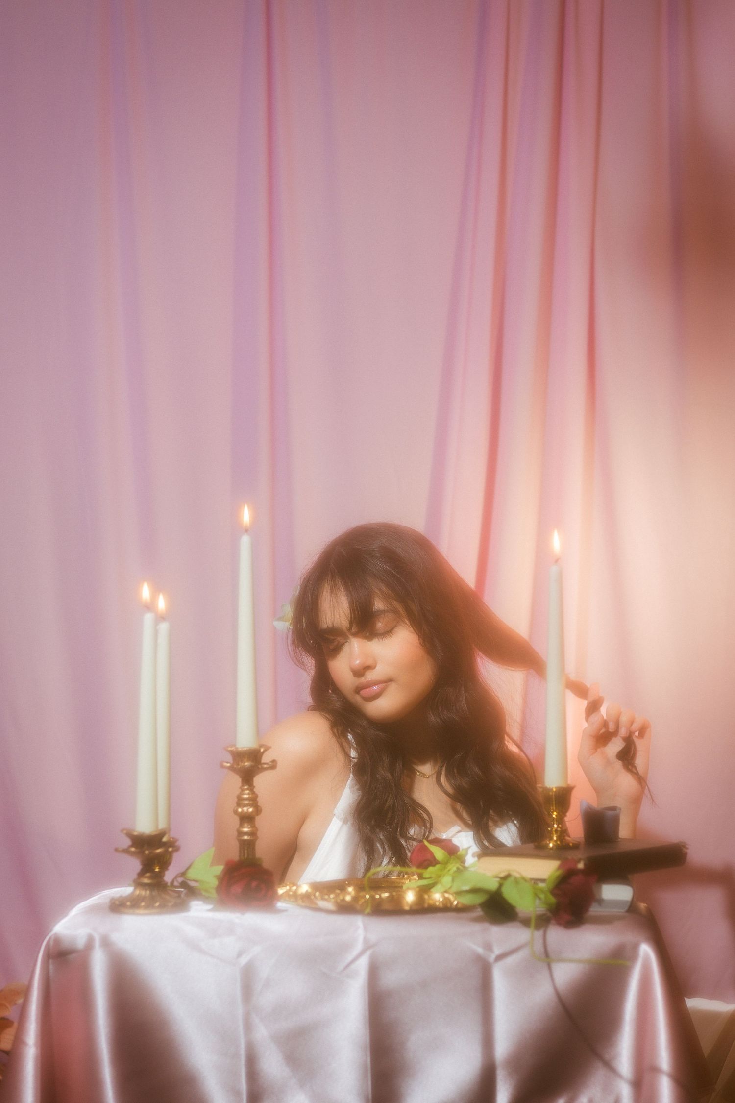 Woman in white dress seated at candlelit table with roses and gold decor against pink drapes.