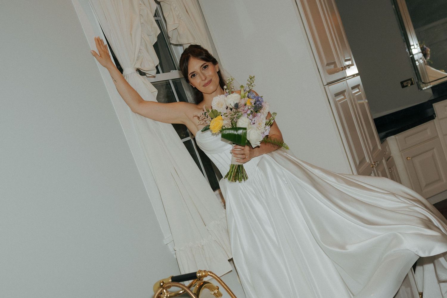 Bride in white gown holding colorful bouquet poses elegantly in a bright room.