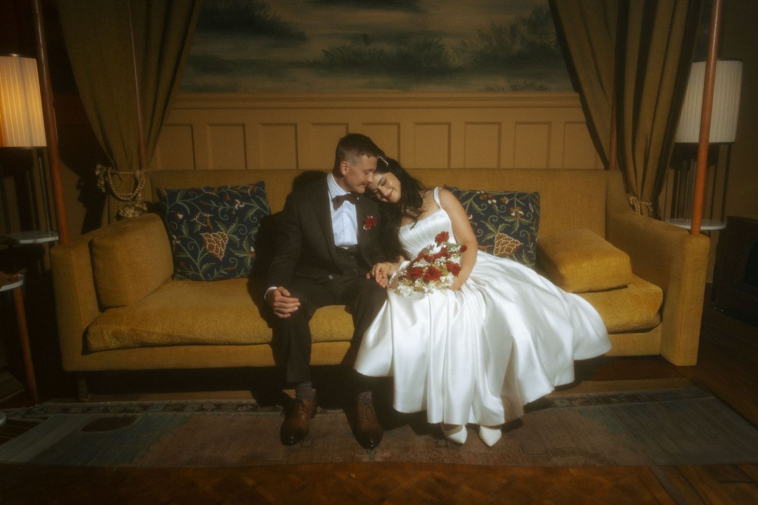 Bride and groom sharing intimate moment on golden sofa, bride laughing while holding red and white bouquet.
