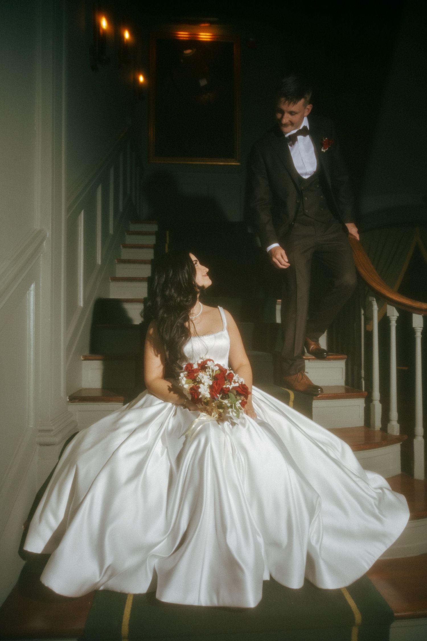 Bride in white satin ballgown sits on staircase holding red bouquet as groom descends in dark suit.