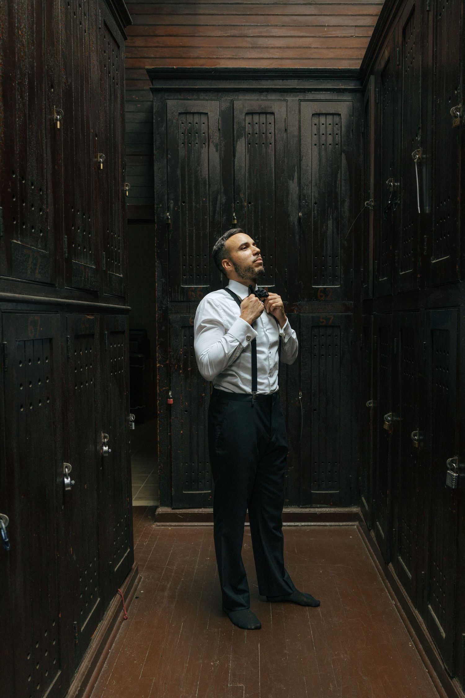 A groom adjusts his tie in a dark wood-paneled locker room before his wedding ceremony.