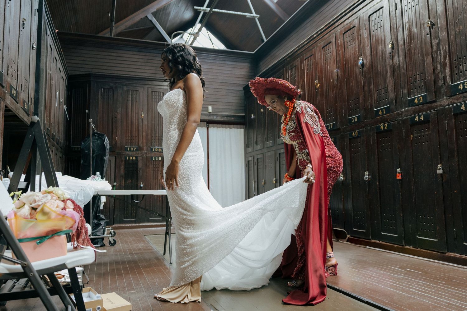 Bride in white gown being helped with dress train in wooden locker room.
