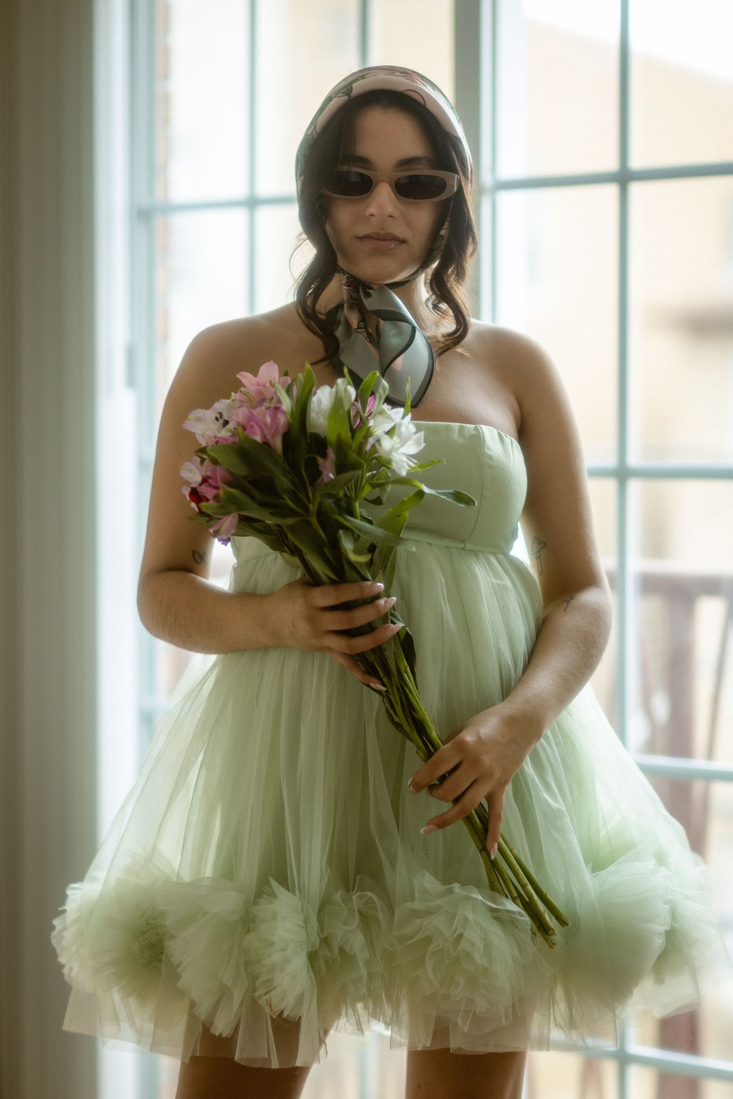 Woman in mint green tulle strapless mini dress holding pink and white flowers, wearing sunglasses and headscarf.