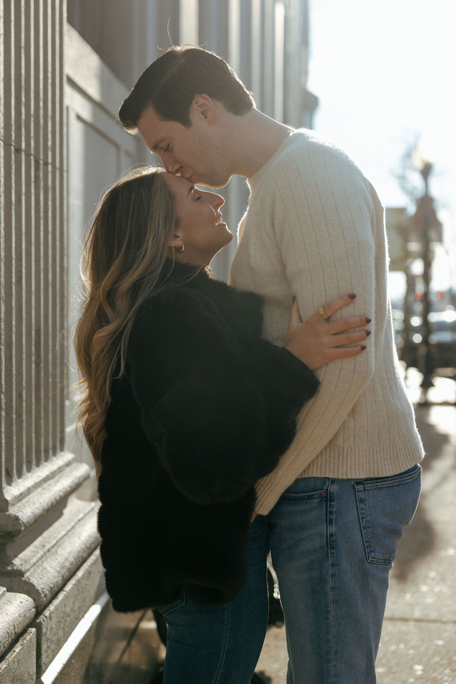 A couple sharing a romantic kiss on a sunny urban sidewalk, woman in black sweater and man in cream knit sweater.