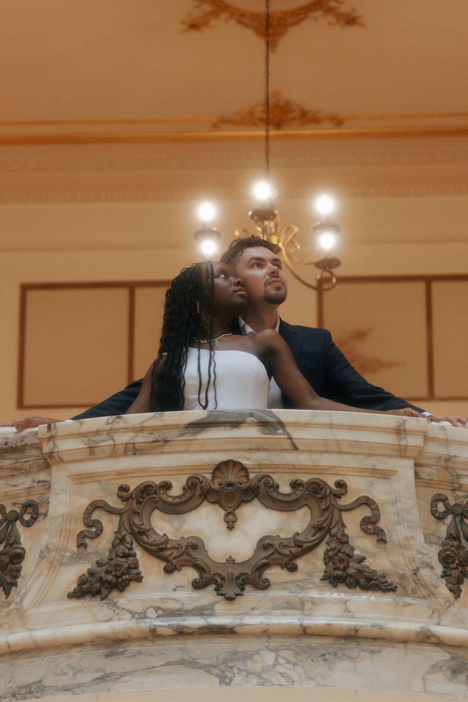 Couple in formal attire embracing on an ornate marble balcony beneath a chandelier in an elegant venue.