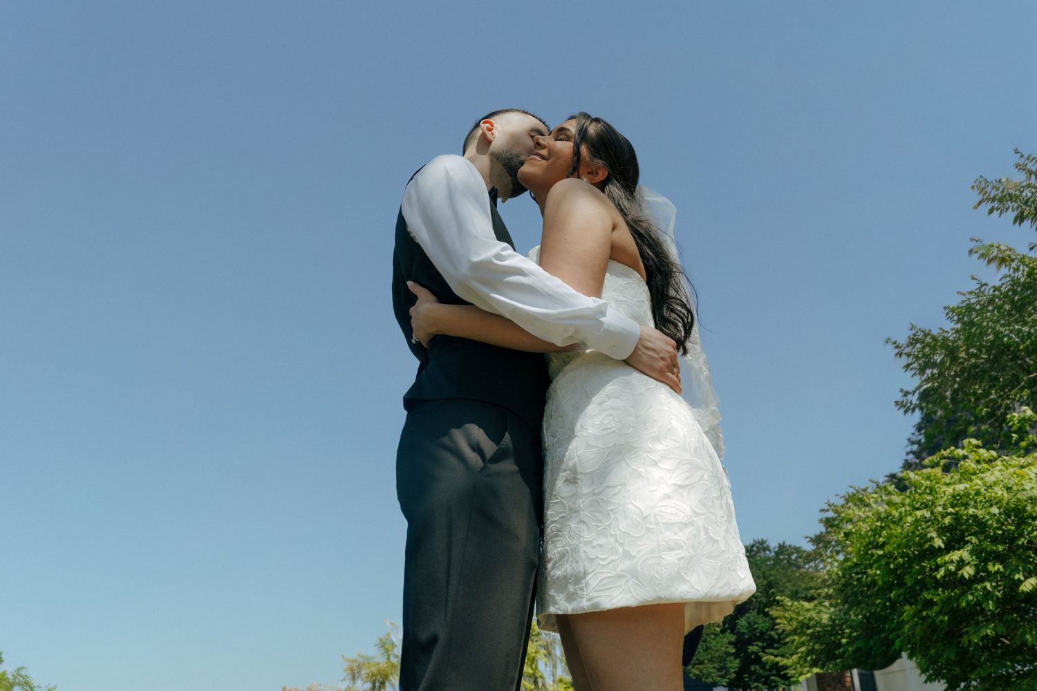 A couple shares a romantic kiss outdoors on their wedding day, bride in white mini dress under a clear blue sky.