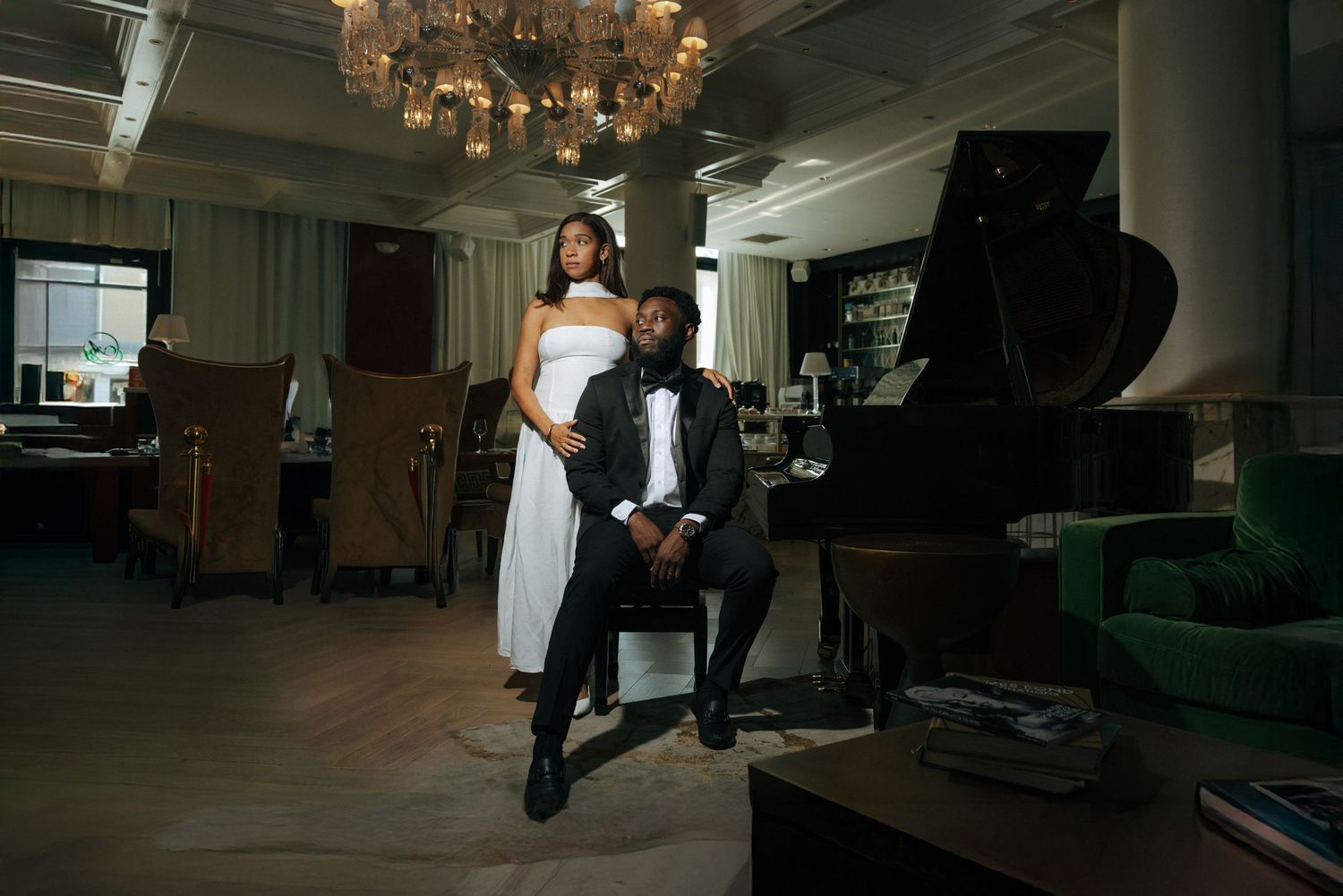 Elegant couple posing near a grand piano in a sophisticated lounge, man in tuxedo and woman in white gown.