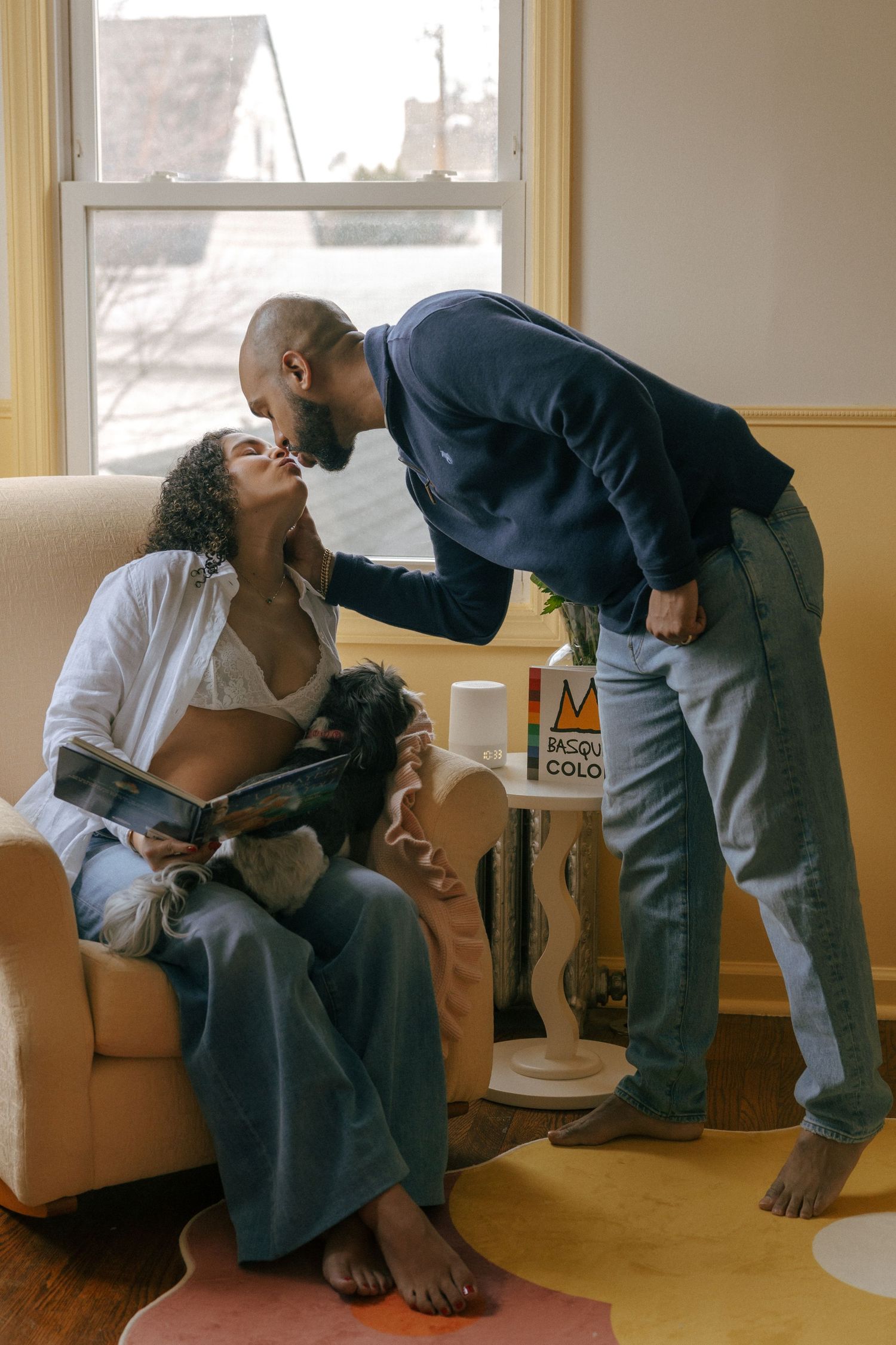 A couple shares a tender kiss, woman seated holding a book and small dog, man leaning in, in a cozy living room.