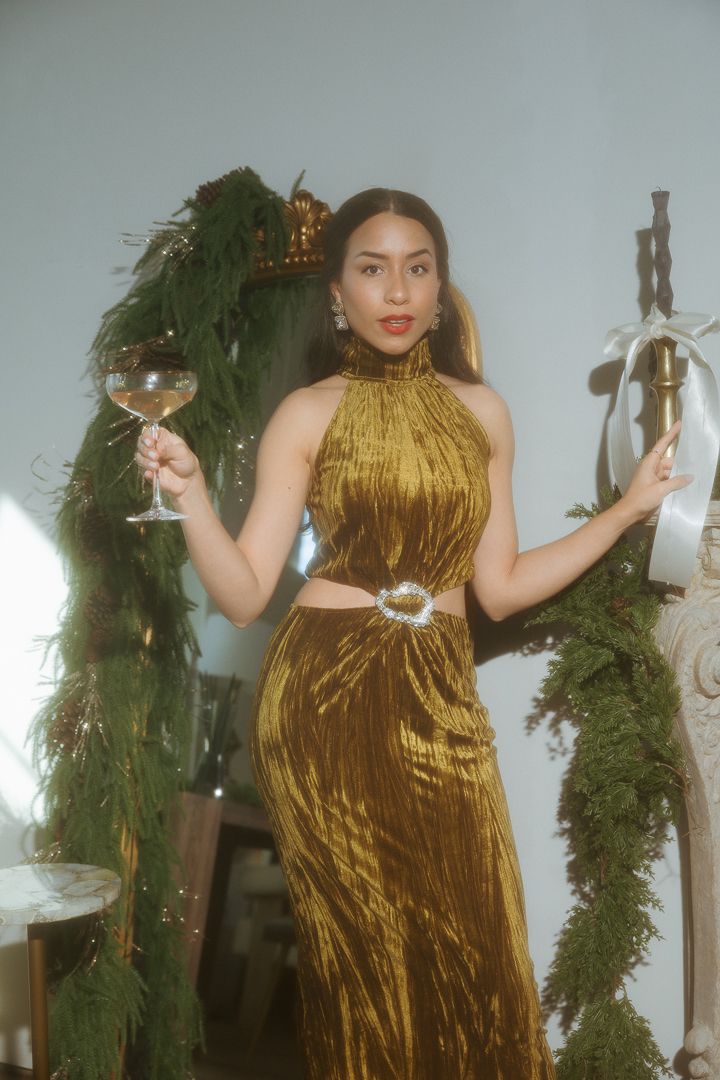 Woman in gold velvet dress raising champagne glass among festive holiday greenery decorations.
