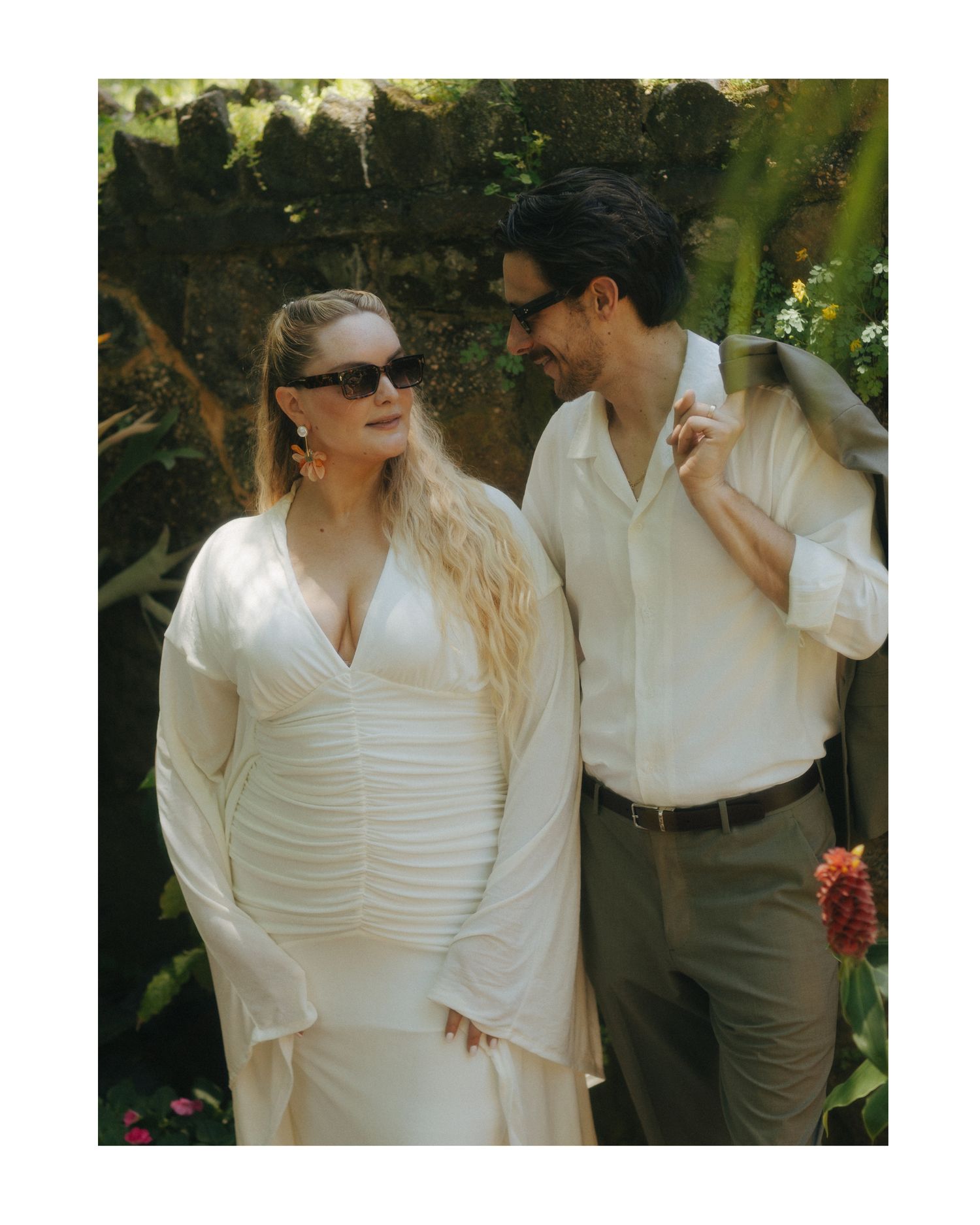 Couple in white outfits with sunglasses posing in a lush garden setting, woman in elegant ruched dress.