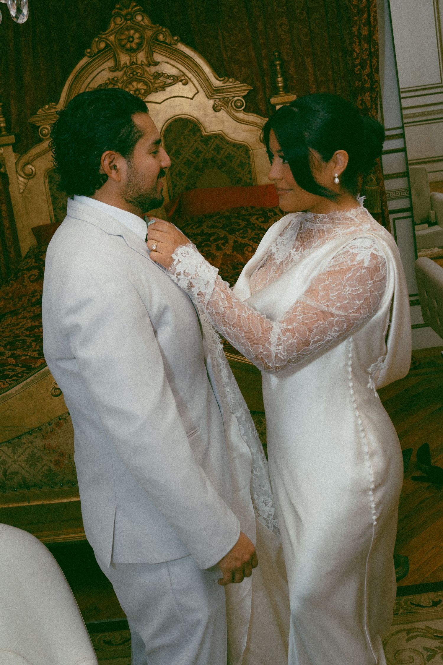 A bride in a lace wedding gown adjusts the groom's white suit collar in an elegant room.