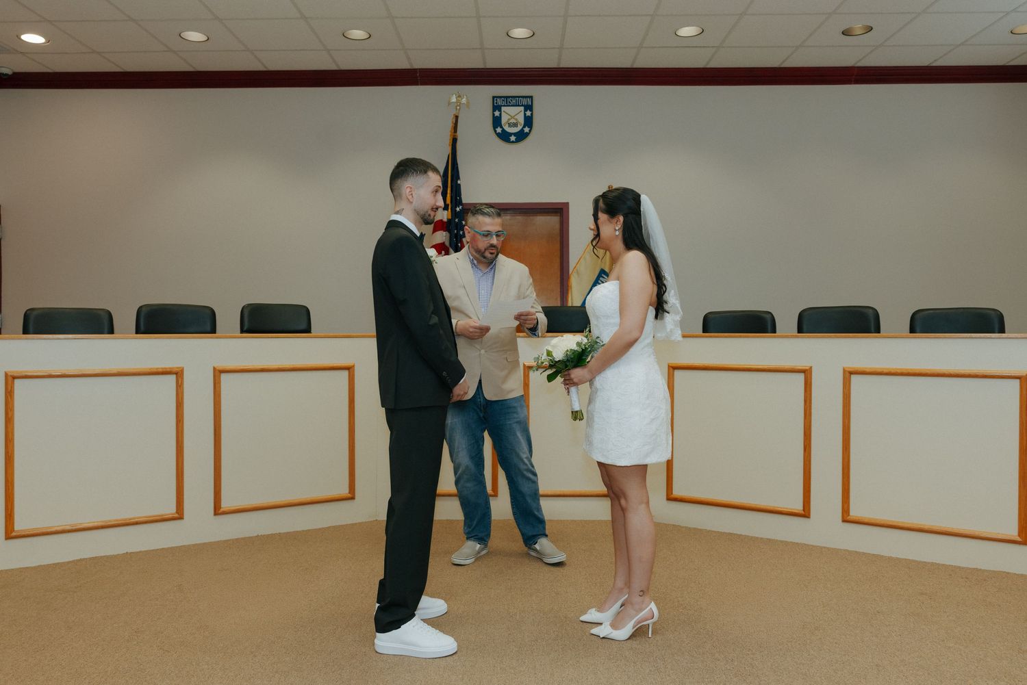 Couple exchanging vows in a courthouse ceremony with an officiant, bride in white mini dress, groom in black suit.