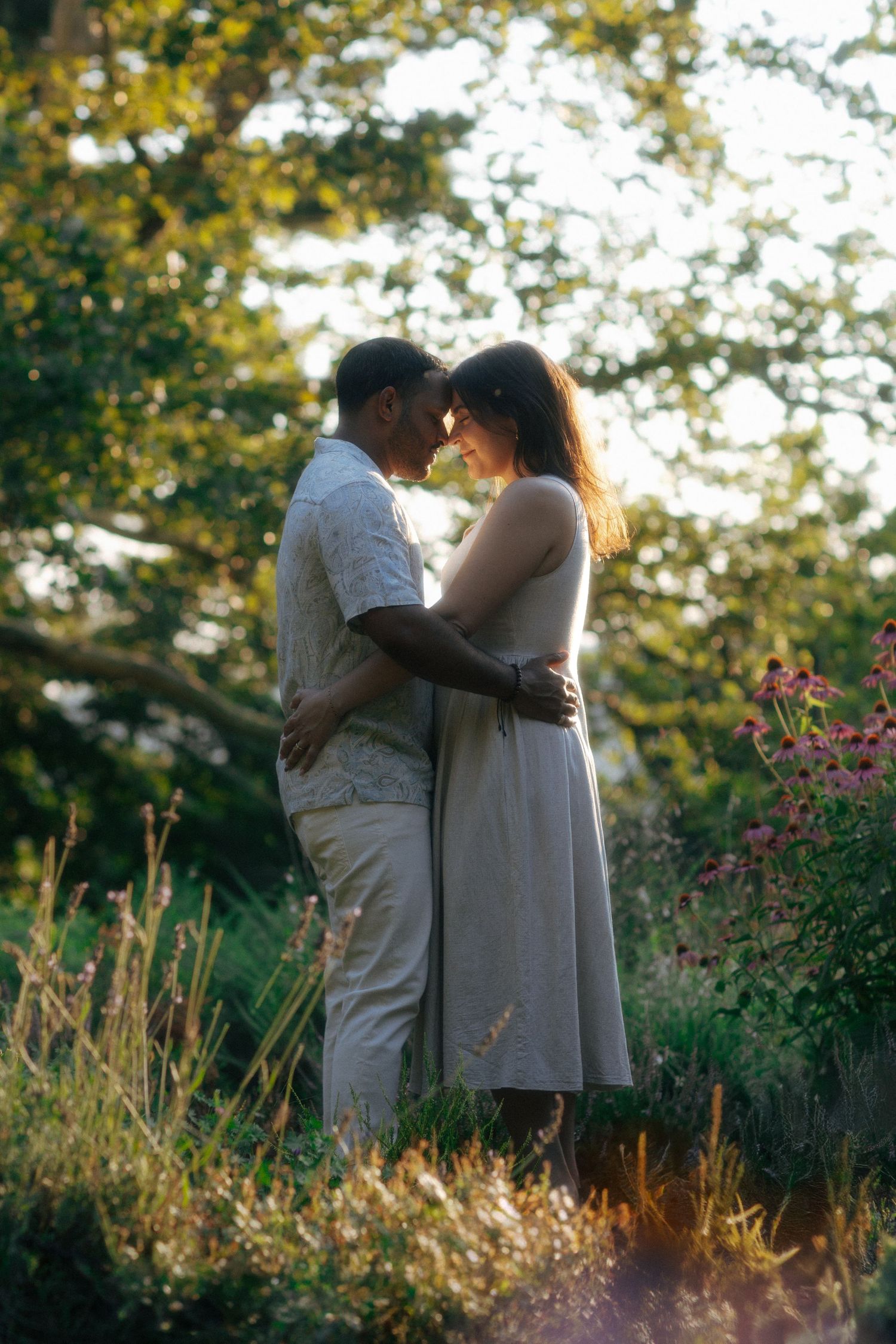 A couple embraces tenderly in a sunlit garden, surrounded by wildflowers and lush green trees.