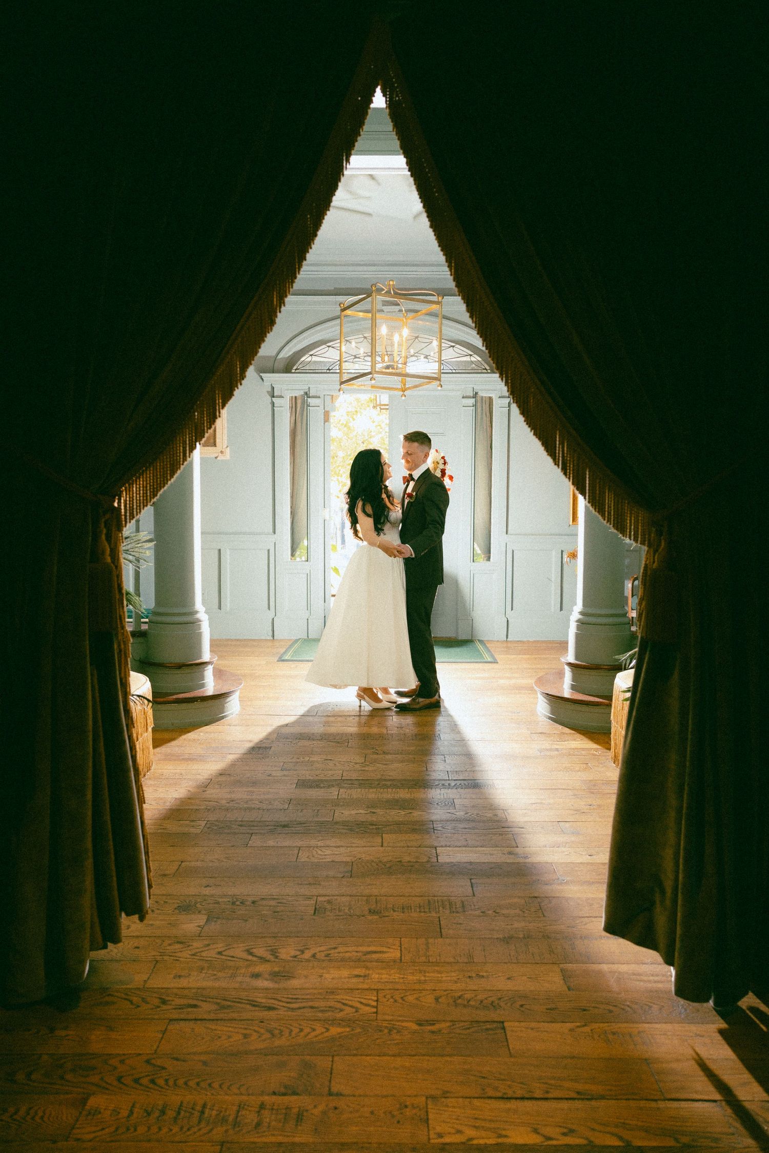 Bride and groom share a romantic moment in an elegant hall with hardwood floors, framed by dark velvet curtains.