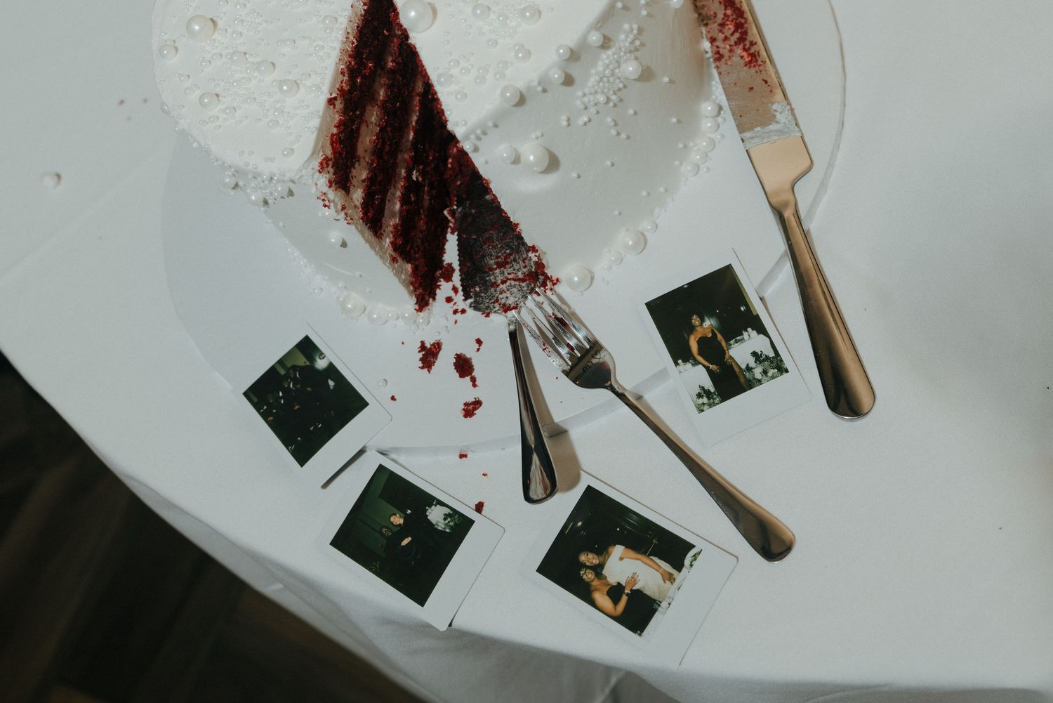 Red velvet wedding cake with white frosting, a slice cut out, fork and knife beside Polaroid photos on a white table.