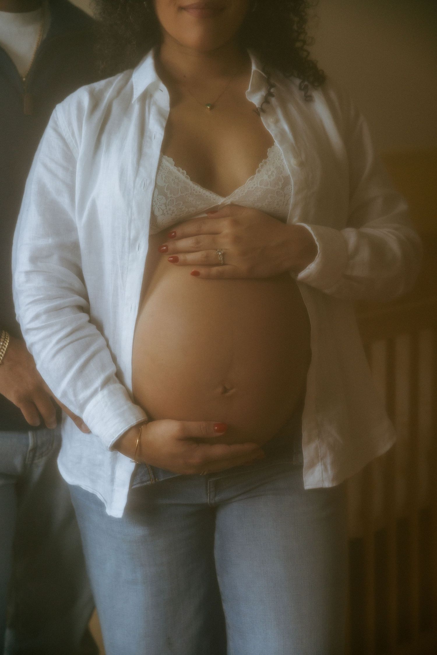 Pregnant woman cradling bare baby bump, wearing open white linen shirt, lace bralette, and blue jeans.