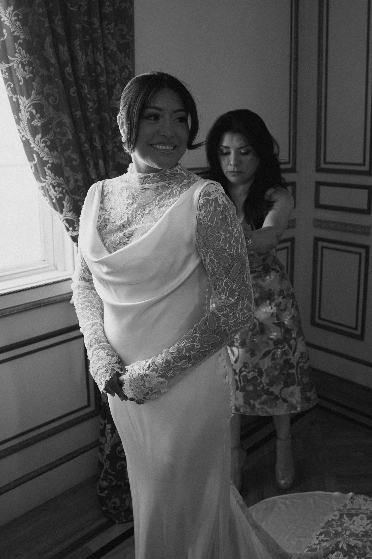 Black and white photo of a bride in lace wedding gown being helped by a woman in a floral dress near a window.