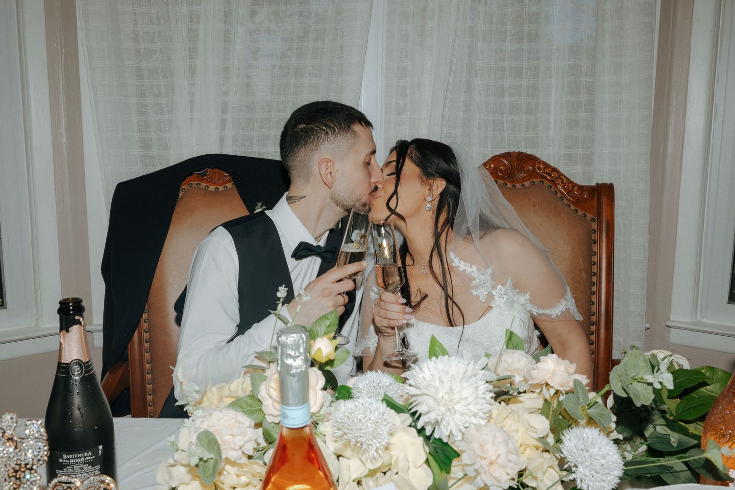 Newlywed couple sharing a kiss while toasting with champagne glasses at their elegant wedding reception table.