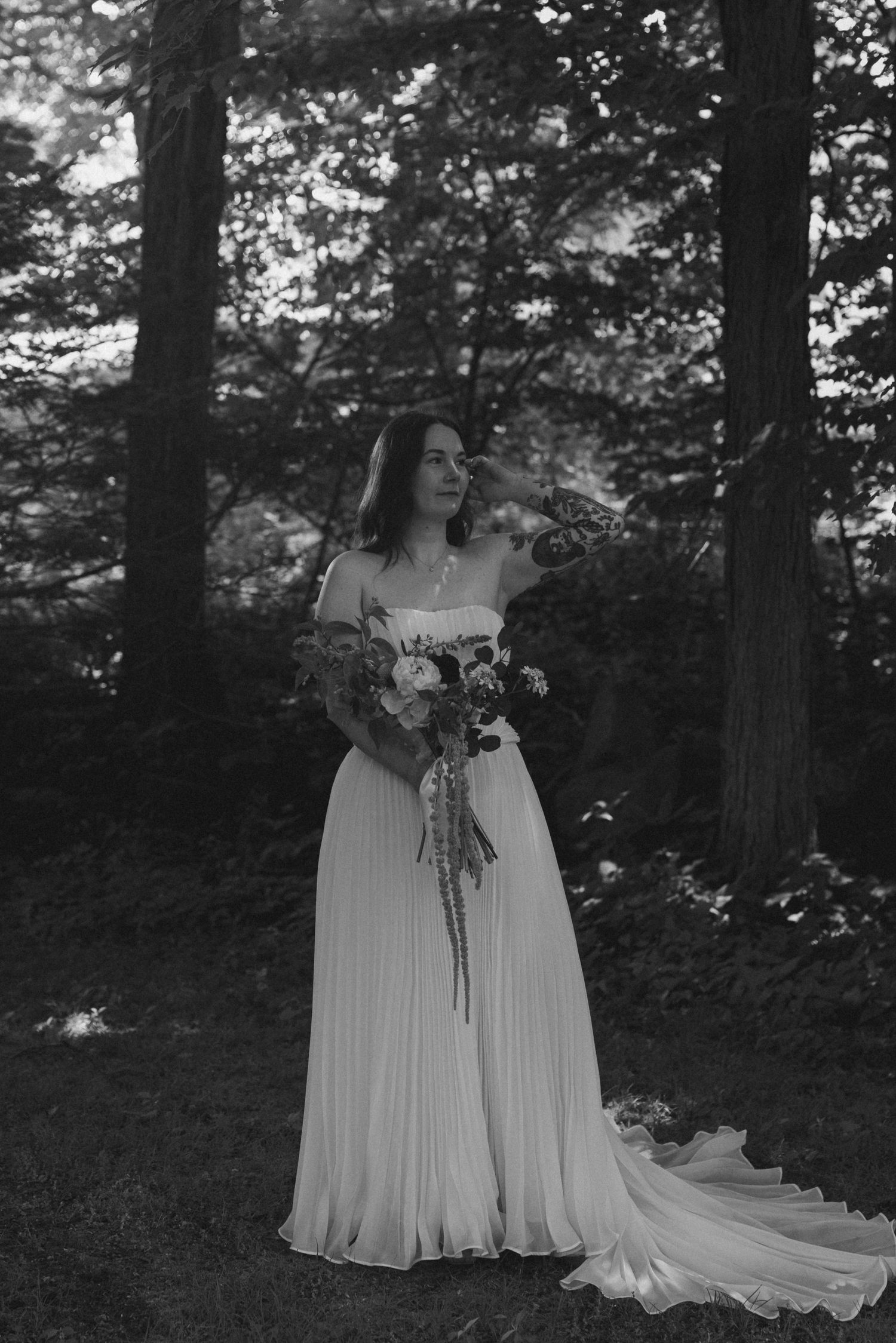 Black and white photo of a bride in a flowing gown holding a bouquet, standing among tall trees in a forest.
