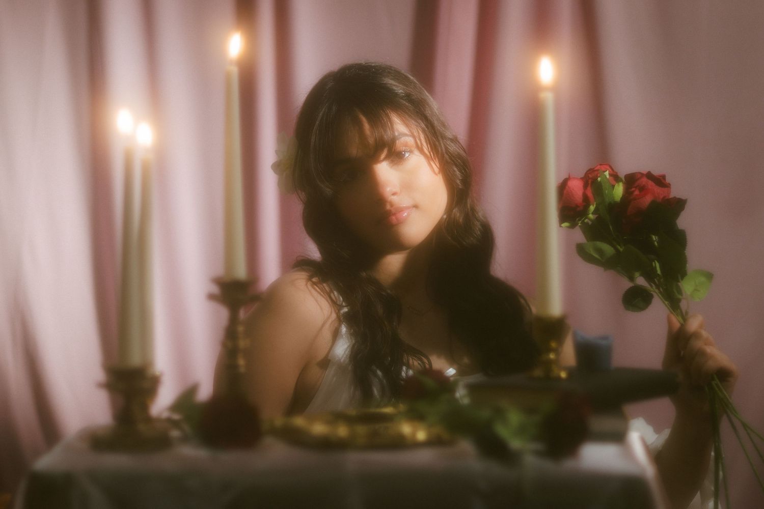 Dreamy portrait of woman holding red roses at candlelit table with soft pink curtain backdrop.