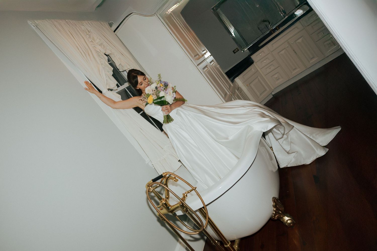 Bride in white wedding dress holding bouquet poses elegantly on ornate gold staircase in luxurious interior.