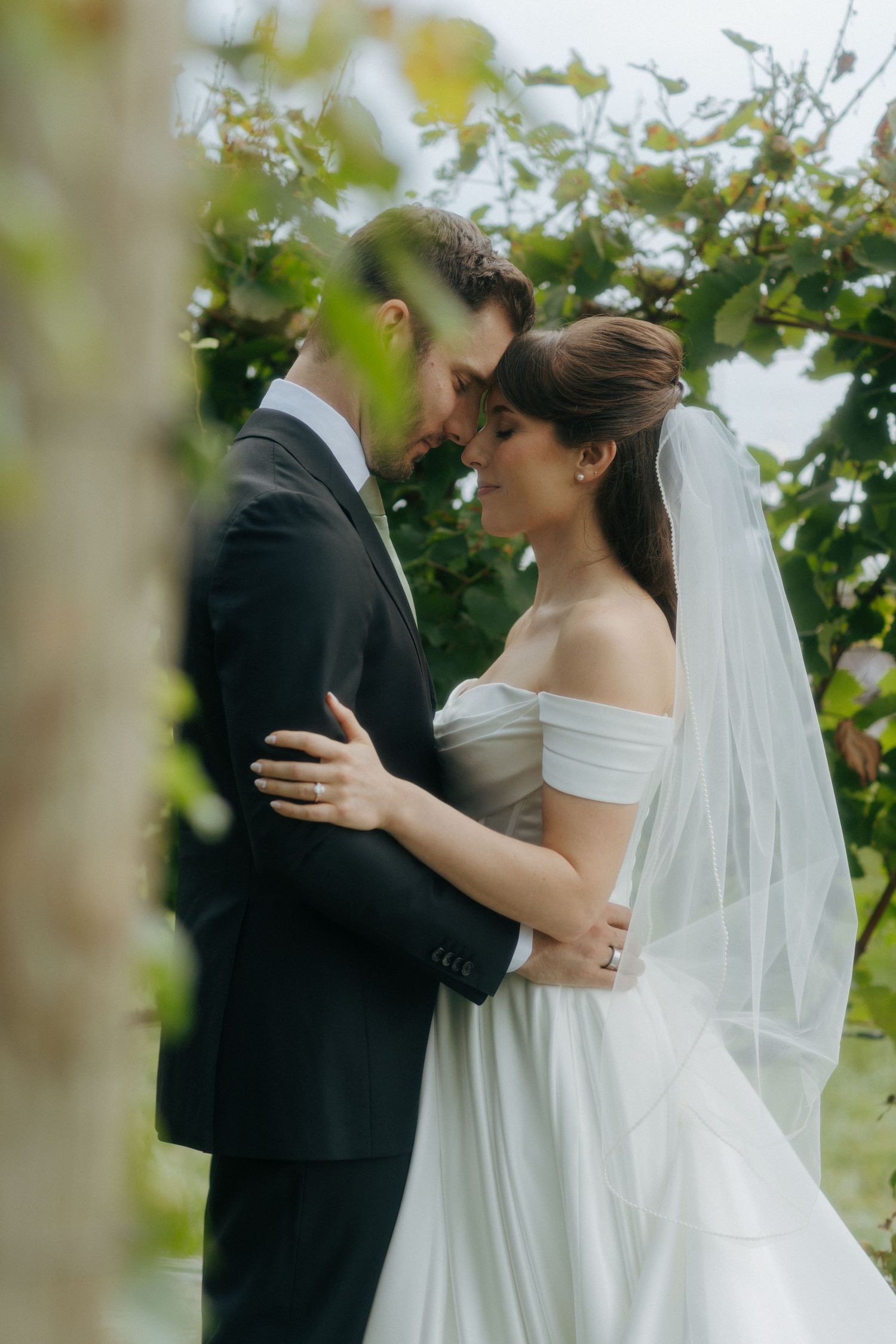 A bride and groom share an intimate moment surrounded by lush greenery, she wearing an off-shoulder gown and veil.