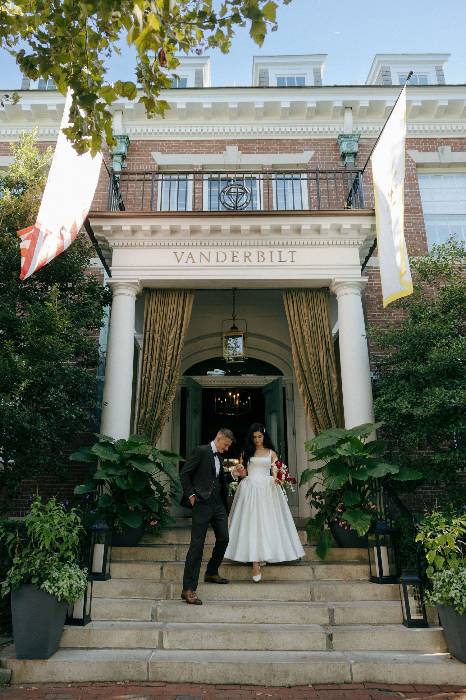 Bride and groom exit the Vanderbilt venue down stone steps surrounded by lush greenery and flags.