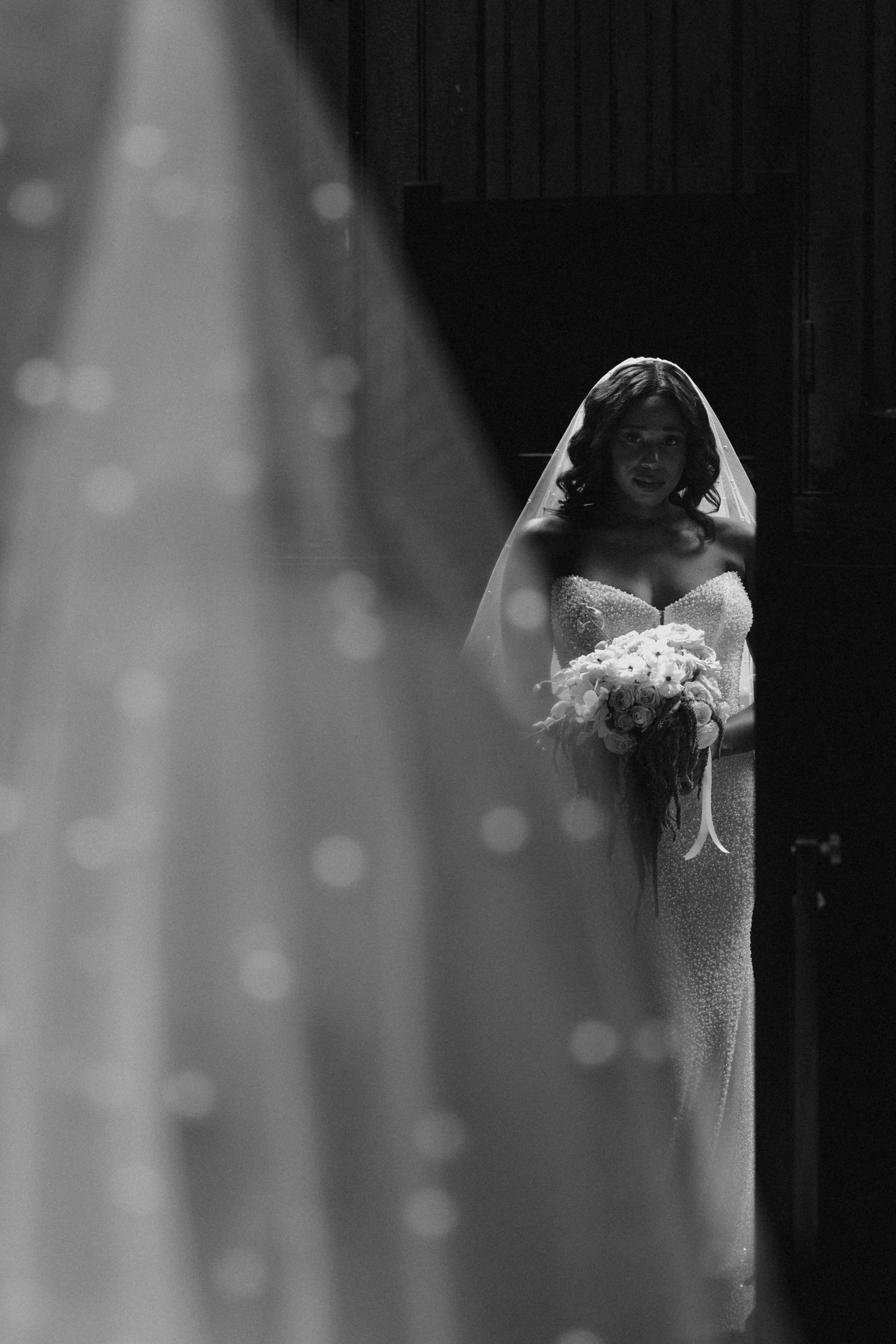Elegant bride in lace gown and veil holds bouquet, captured in stunning black and white wedding portrait.
