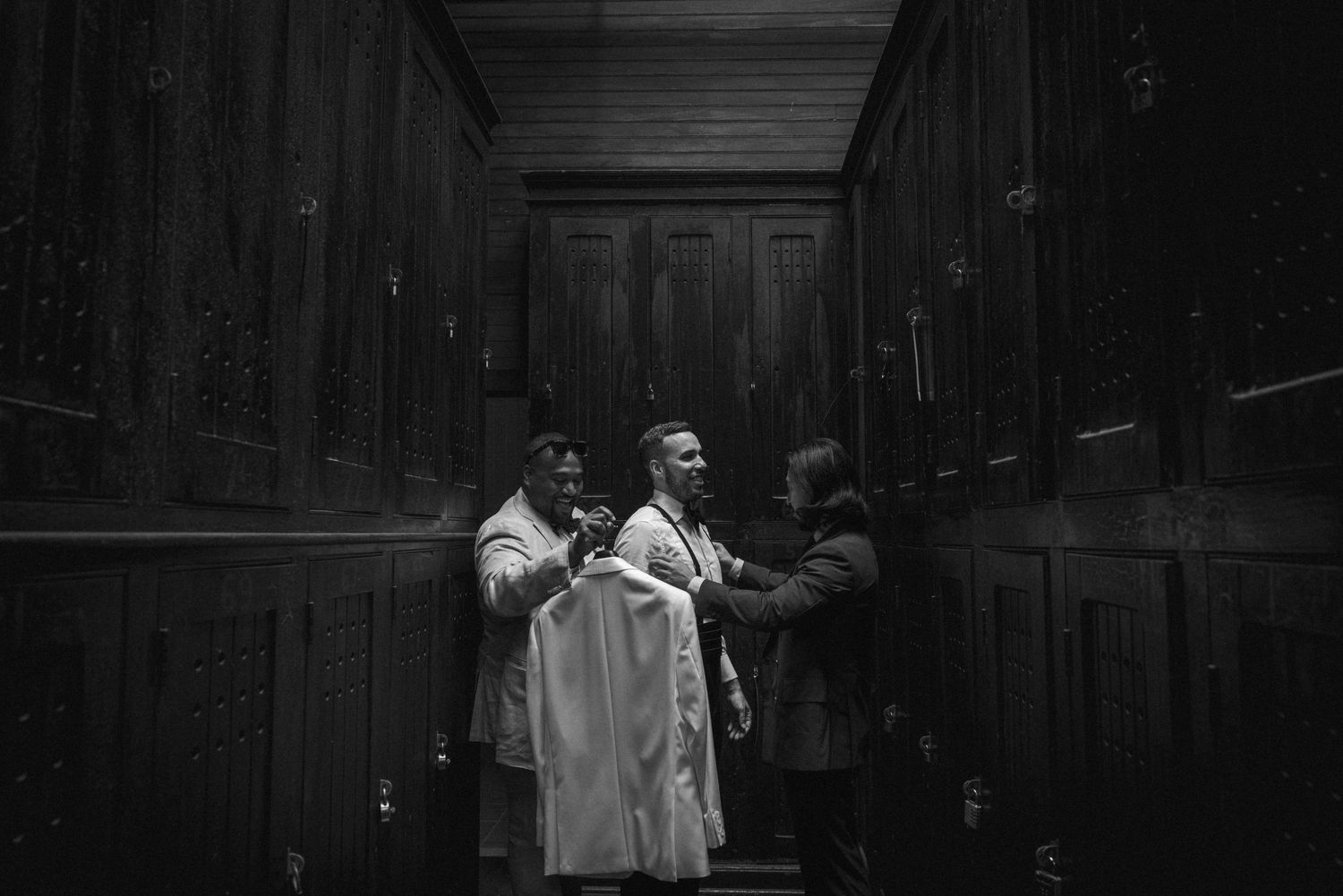Black and white photo of men in formal wear adjusting a white shirt in a dark wood-paneled hallway.