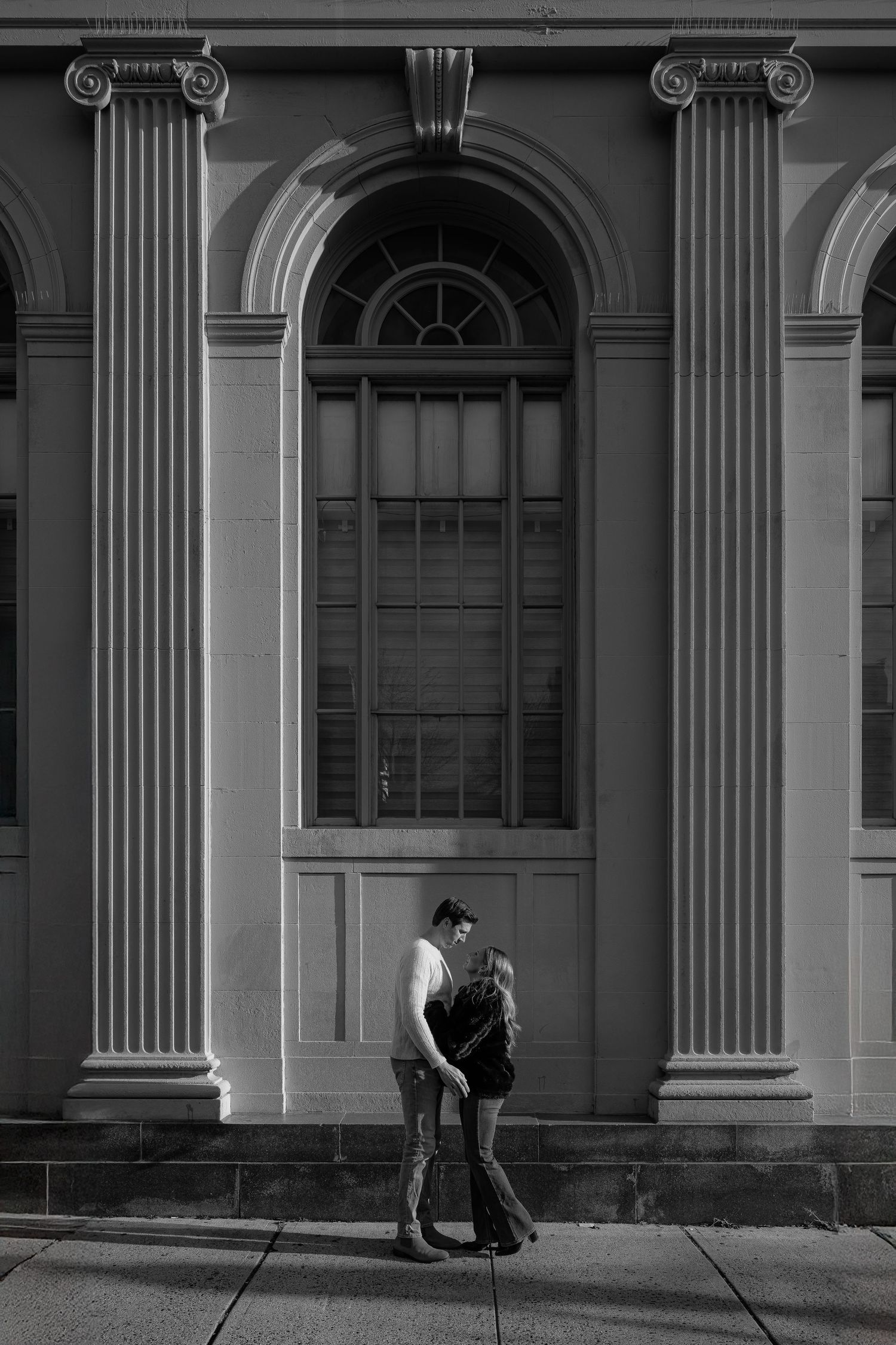 Black and white photo of a couple embracing in front of a classic arched building with ornate columns.