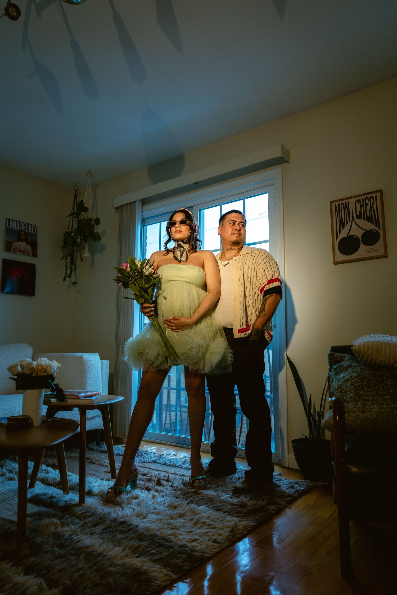 Pregnant woman in tulle skirt holding flowers stands with partner in warmly lit living room.