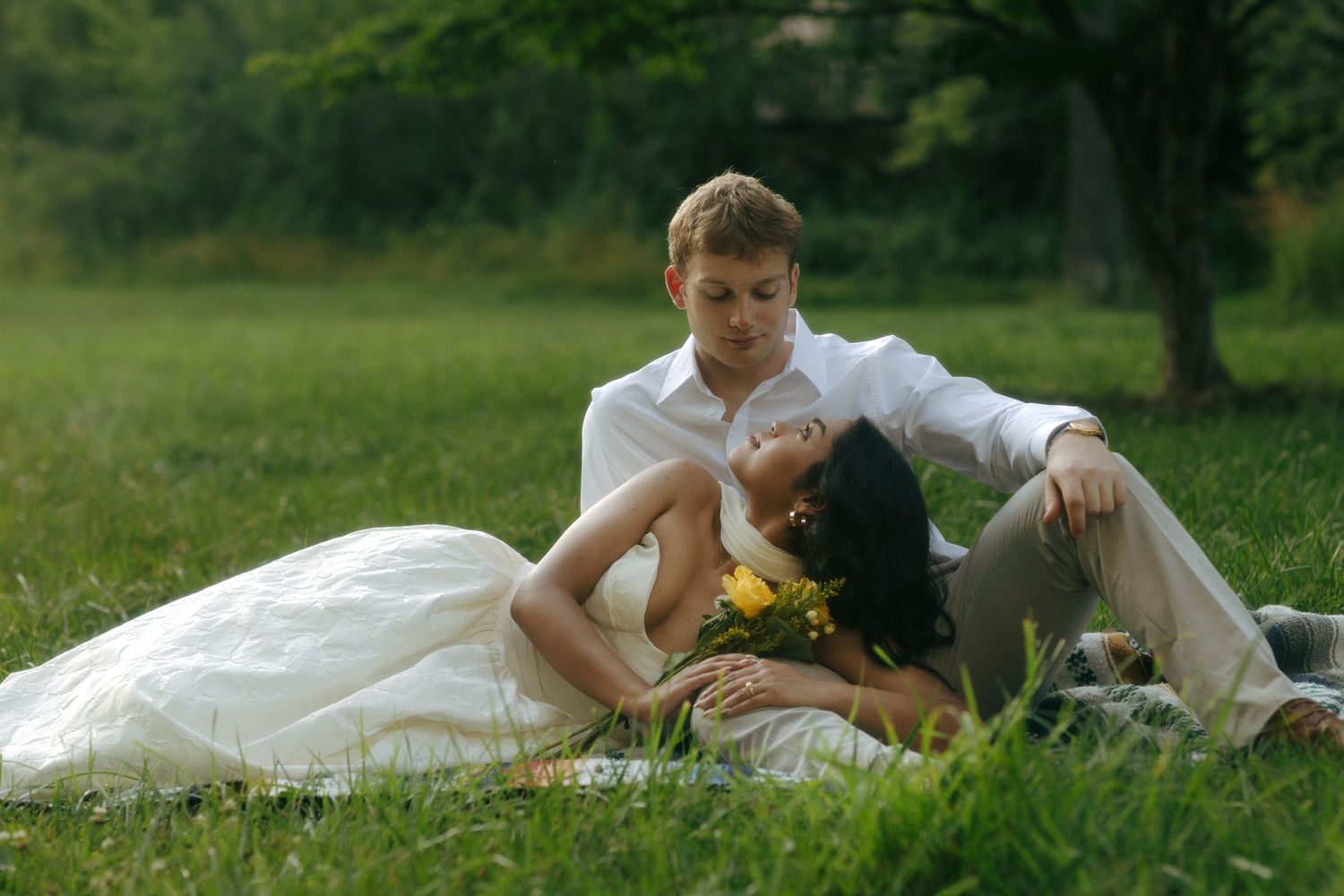 Bride and groom sitting on green grass outdoors, woman in white dress holding yellow flowers, romantic wedding photo.