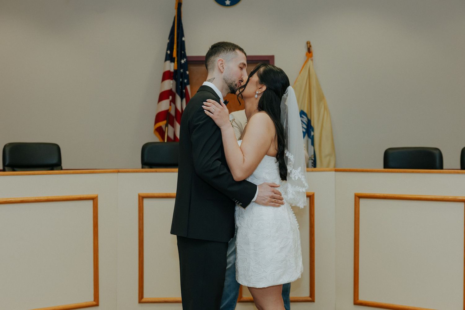 Couple shares first kiss as newlyweds at courthouse wedding ceremony with American flag in background.