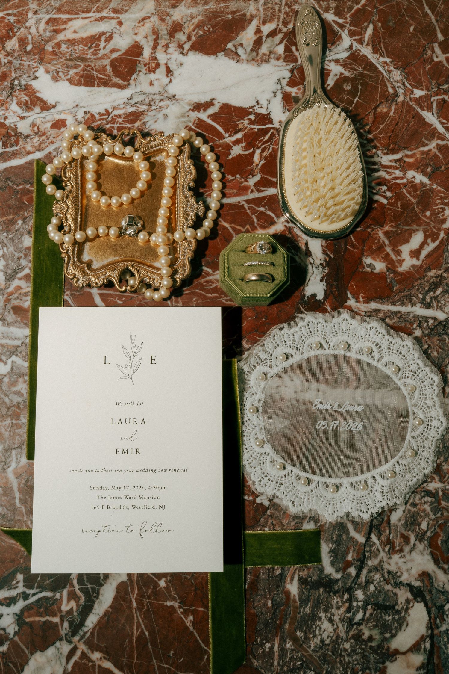 Wedding flat lay with invitation for Laura and Emir, pearl jewelry, gold trinket box, green brush, and rings on marble.